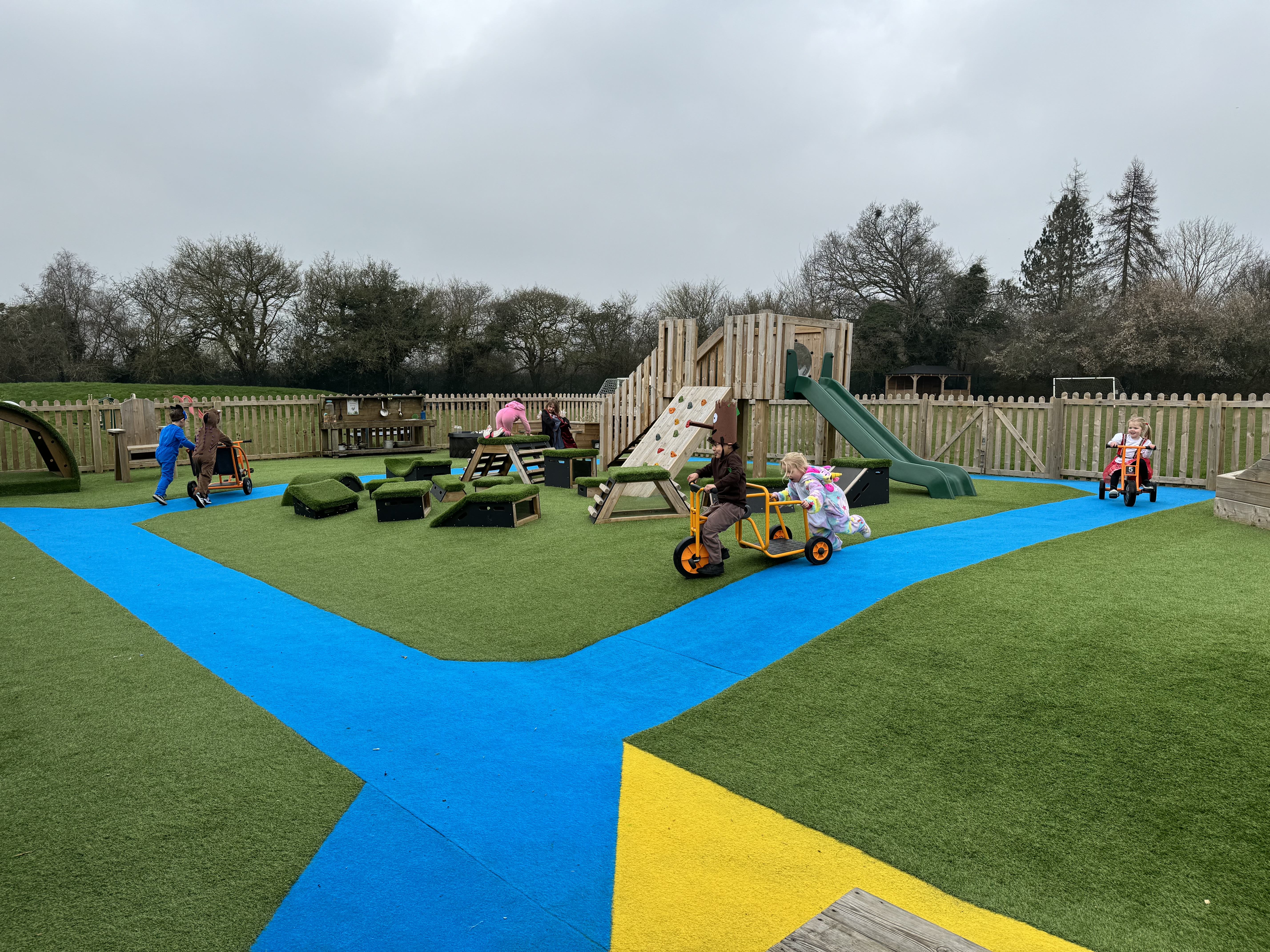 A playground which has artificial grass surfacing in blue, yellow and green colours. A variety of play equipment can be seen with children playing on it.