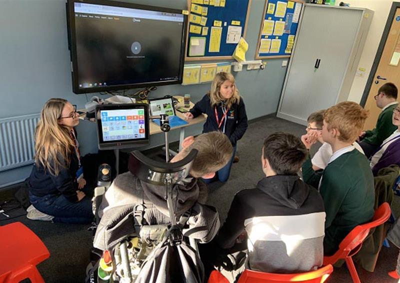 Two Pentagon Play employees are giving a presentation to a group of children regarding playgrounds. The children are all sat on chairs and are watching