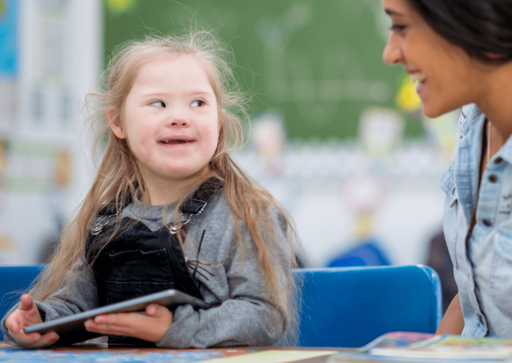 An SEN child is holding an iPad and is looking at a female teaching assistant. The child is smiling as she looks at the teaching assistant.