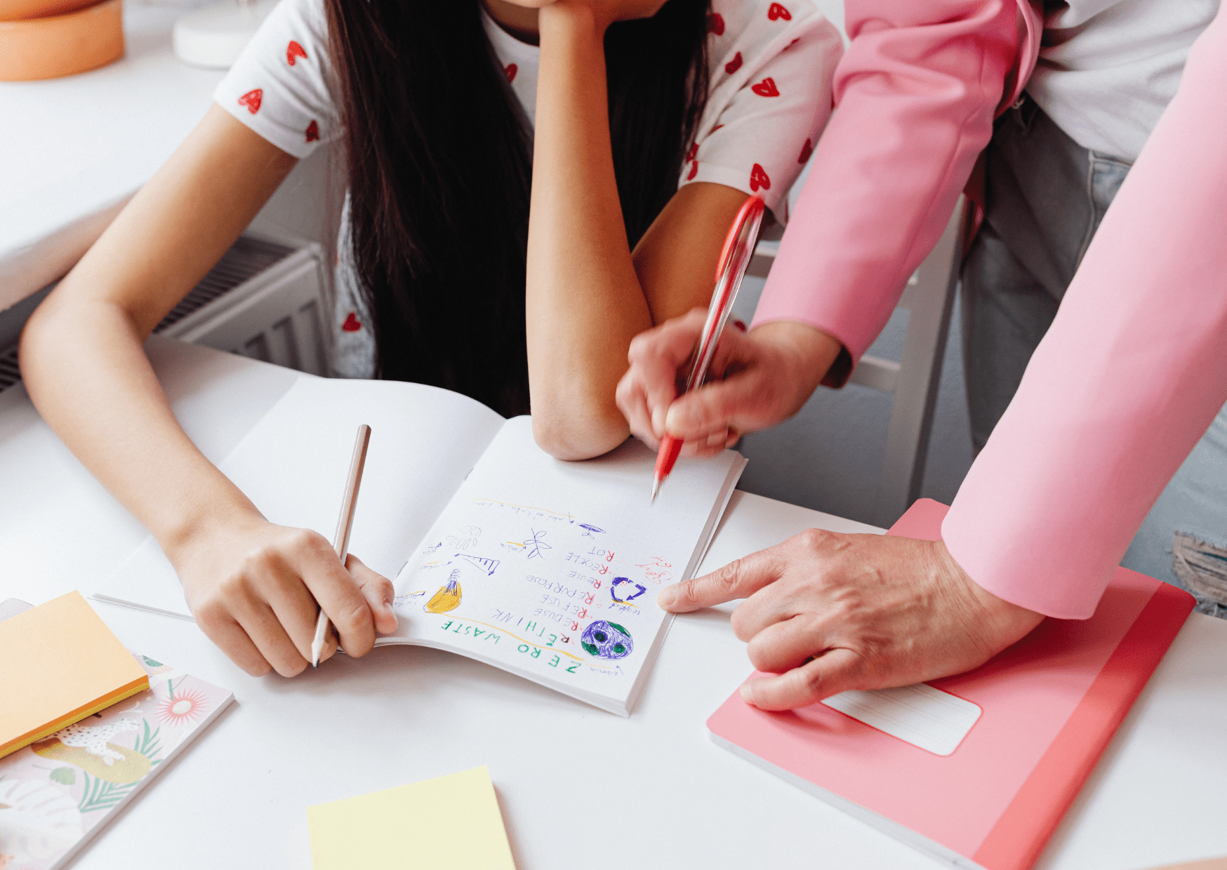 A teacher is helping a child with their work. The notebook is showing writing that surrounds the topic of zero waste. The girl is watching the teacher, who is pointing their pen at specific sentences.