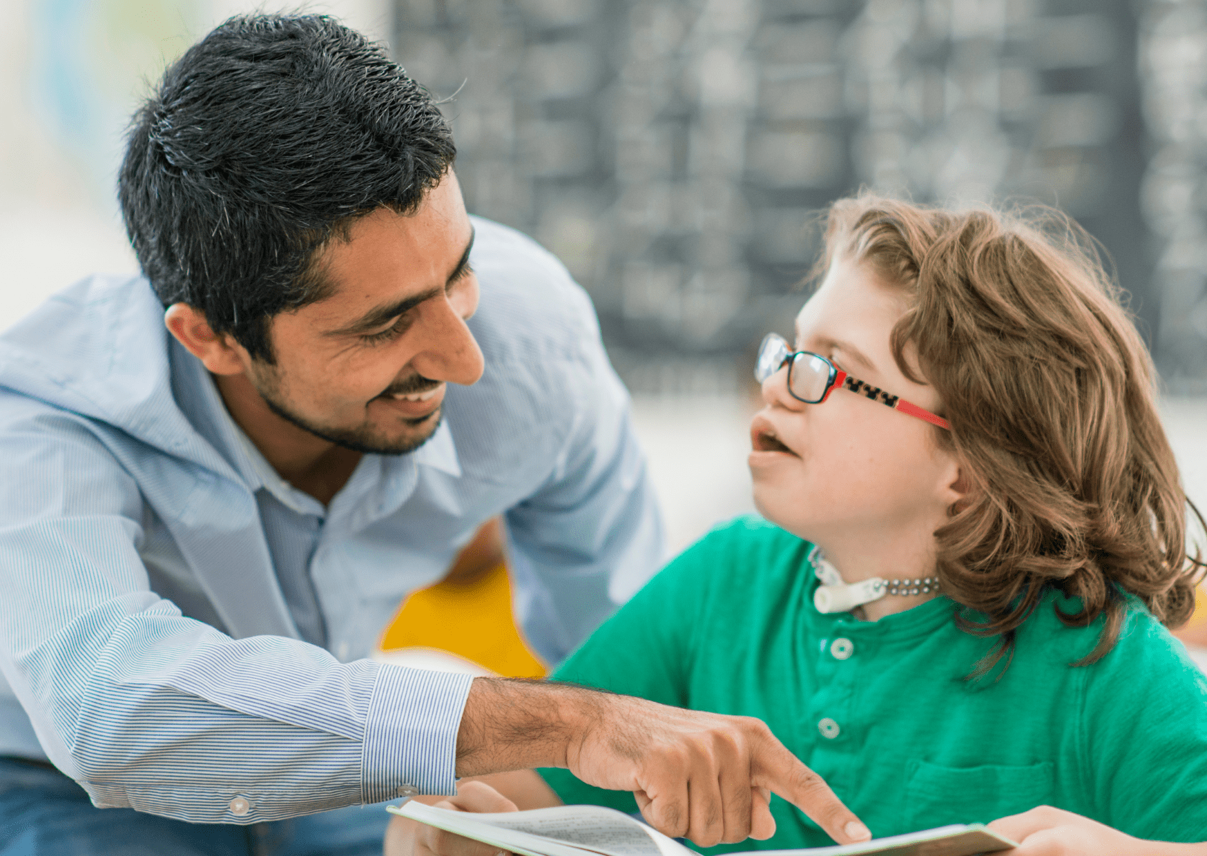 A teaching assistant is smiling whilst helping a little girl with special educational needs. The girl is smiling back to the man.