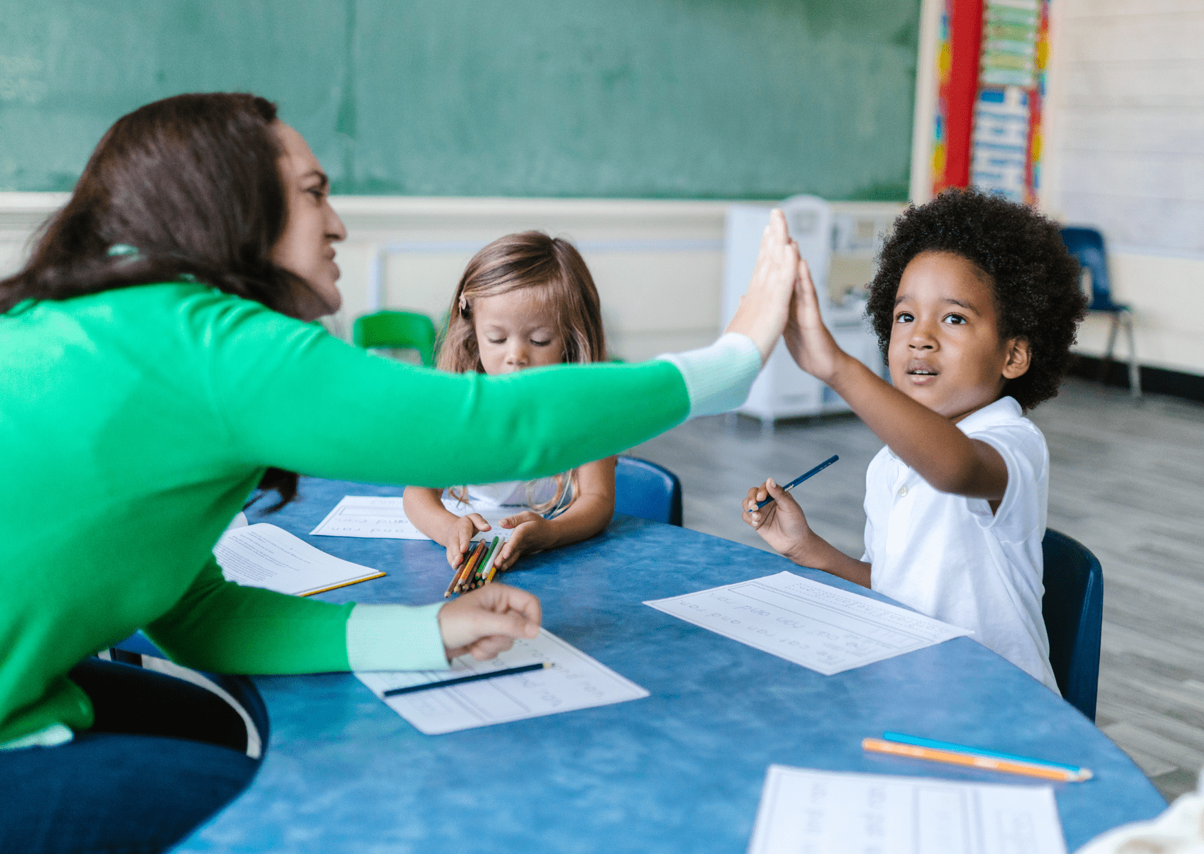 A teaching assistant is giving a young boy a high five after completing a worksheet.