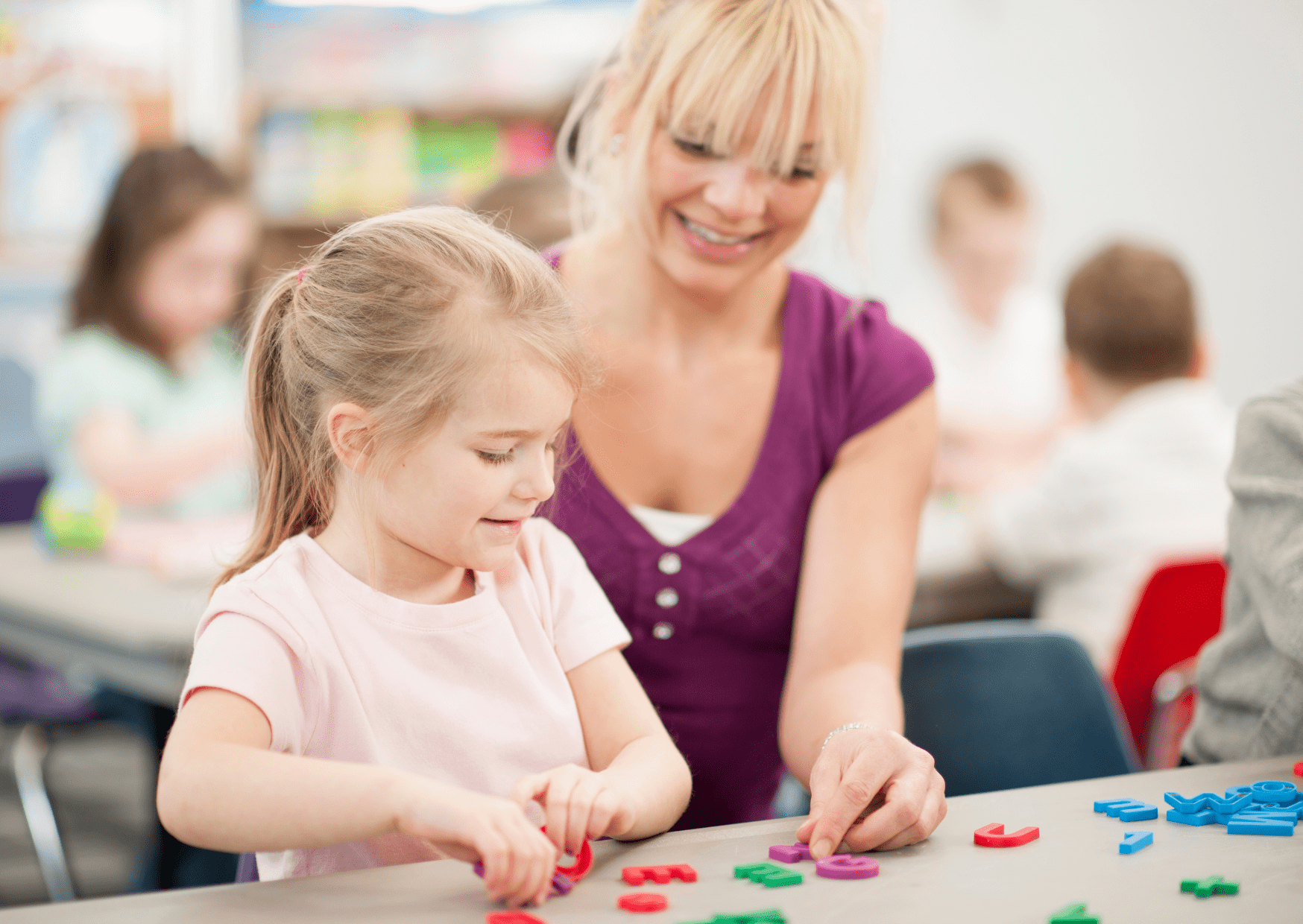 A teaching assistant is sat with a little girl and is helping the girl complete a school task.