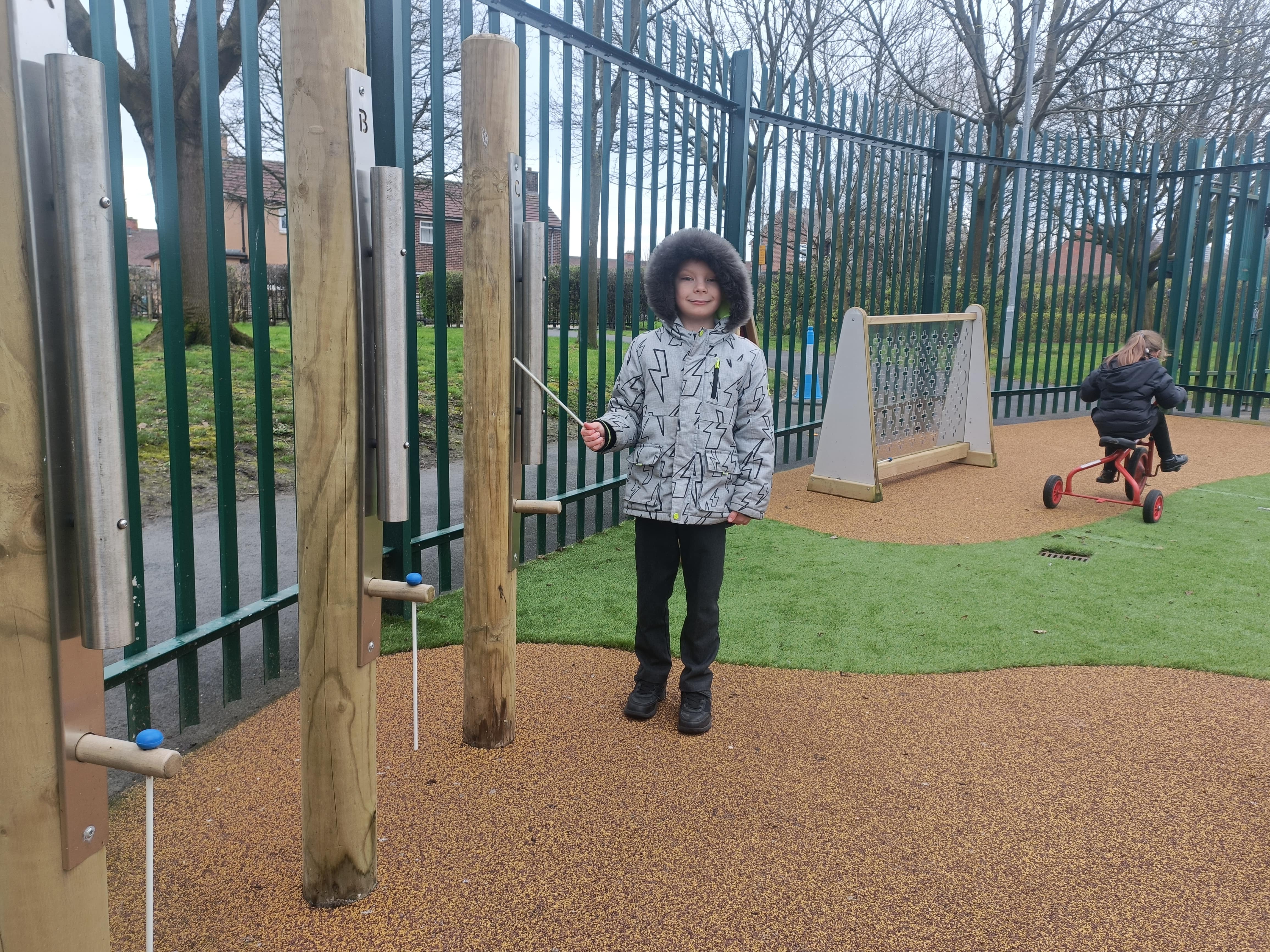 A child is holding a small, soft mallet and is standing next to a variety of different freestanding singular chimes. The child is about to hit one of the chimes. The child and chimes are on artificial grass.