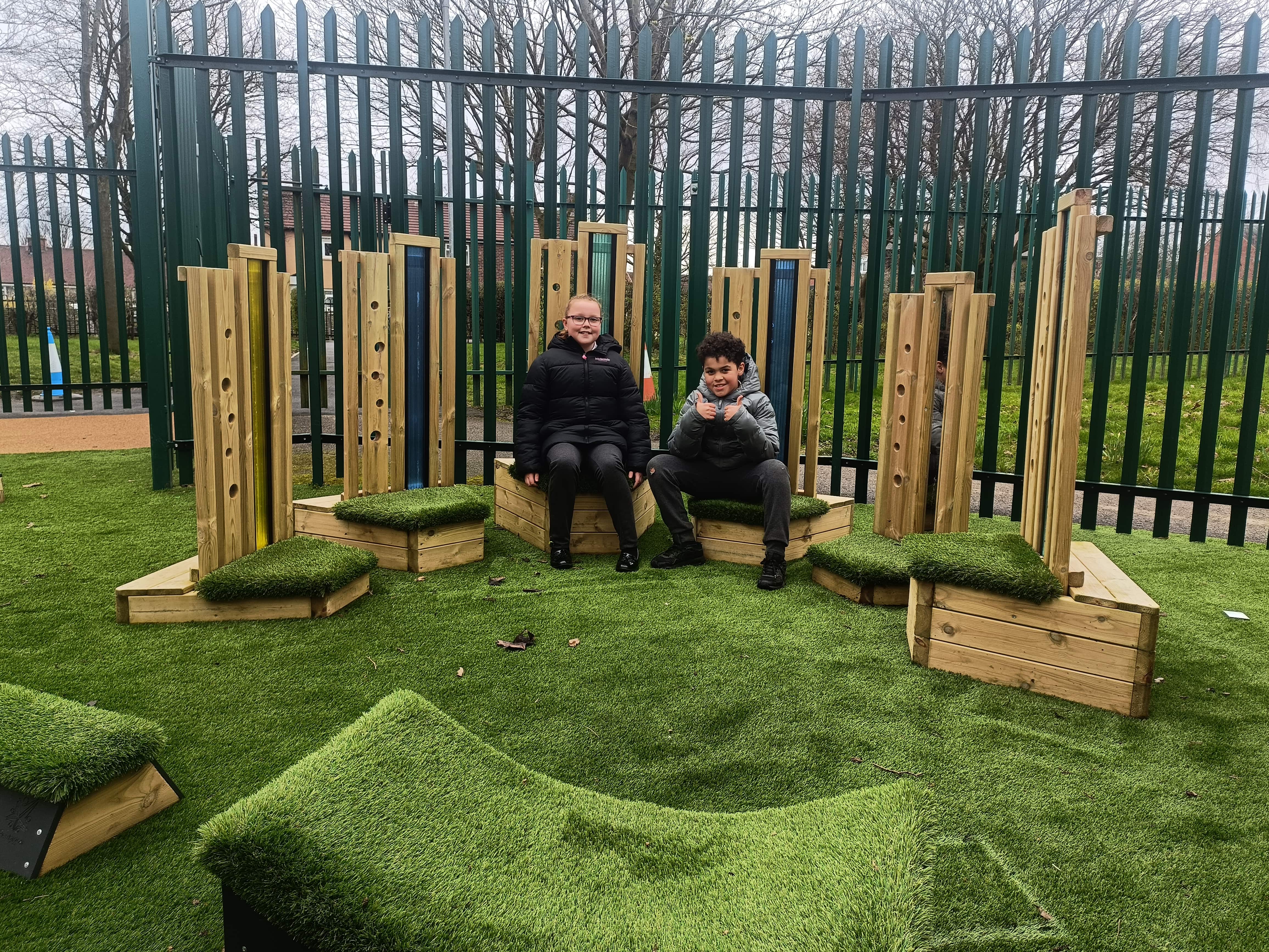 Two children are sat on a seat from the Sensory Seating Circle. The chairs all have long backs, with a different coloured plastic in each back. Each seat is topped with artificial grass. The seats have been placed on top of synthetic turf surfacing.