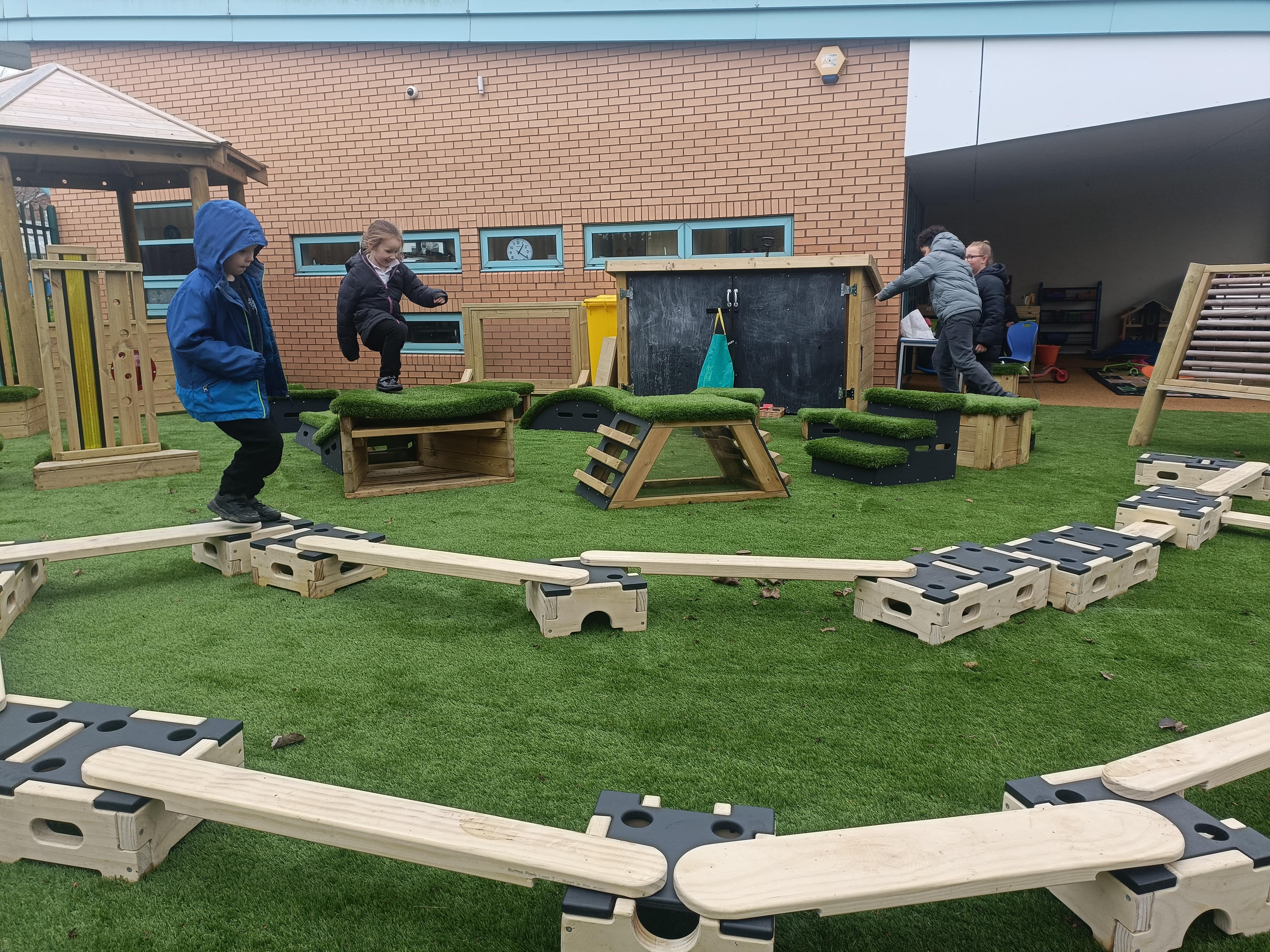 A little child is balancing their way across a play builder set, whilst another group of children are playing on the Get Set, Go! Blocks in the background. All of this is taking place on artificial grass surfacing.