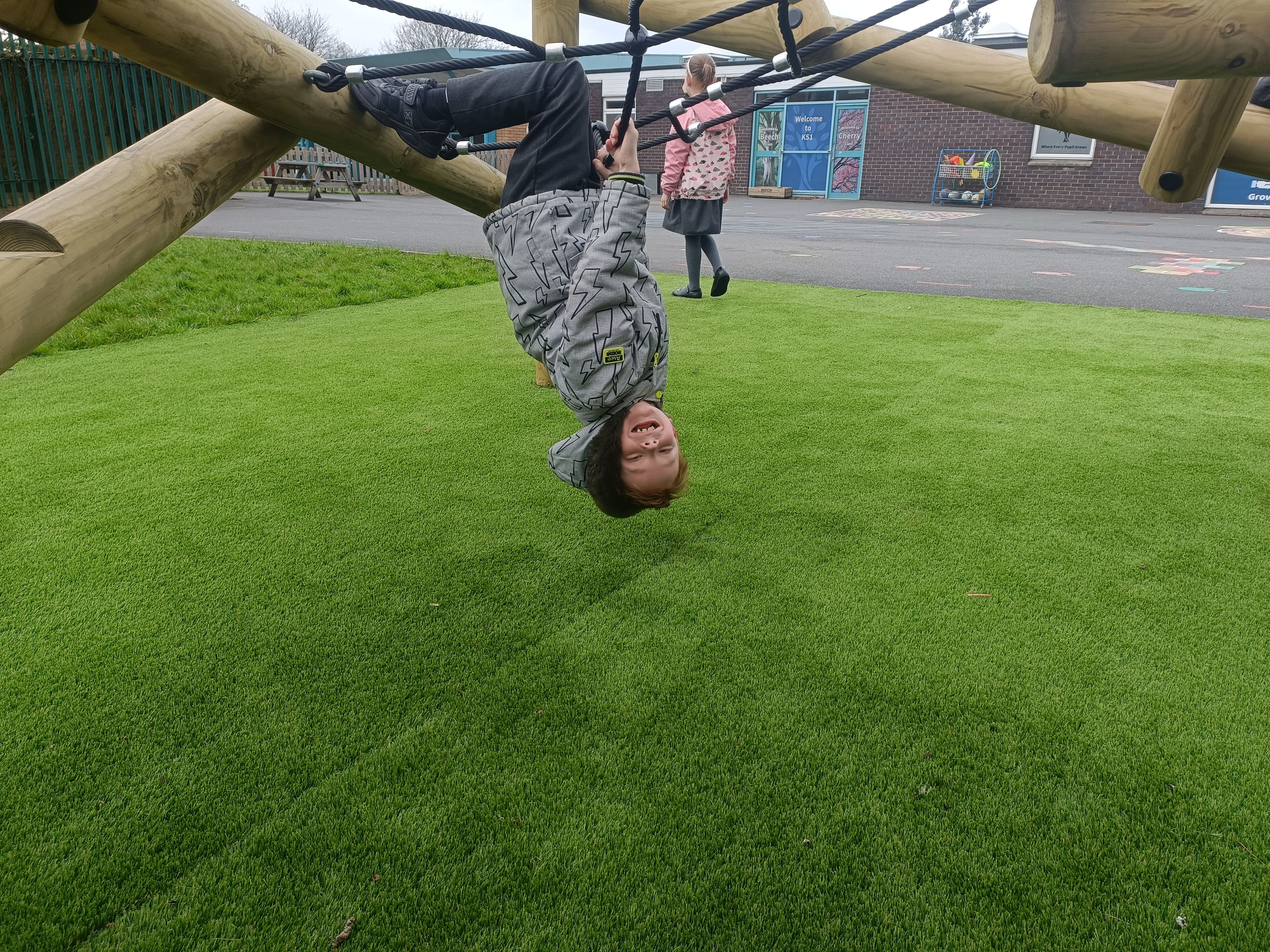 A child is hanging upside down from a climbing frame by holding onto the ropes for support. Beneath the child is a patch of synthetic turf. The child is smiling and happy.