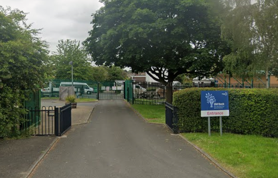 The outside of Old Bank Academy, showcasing a tarmac driveway with a blue little sign post displaying the name of the academy.