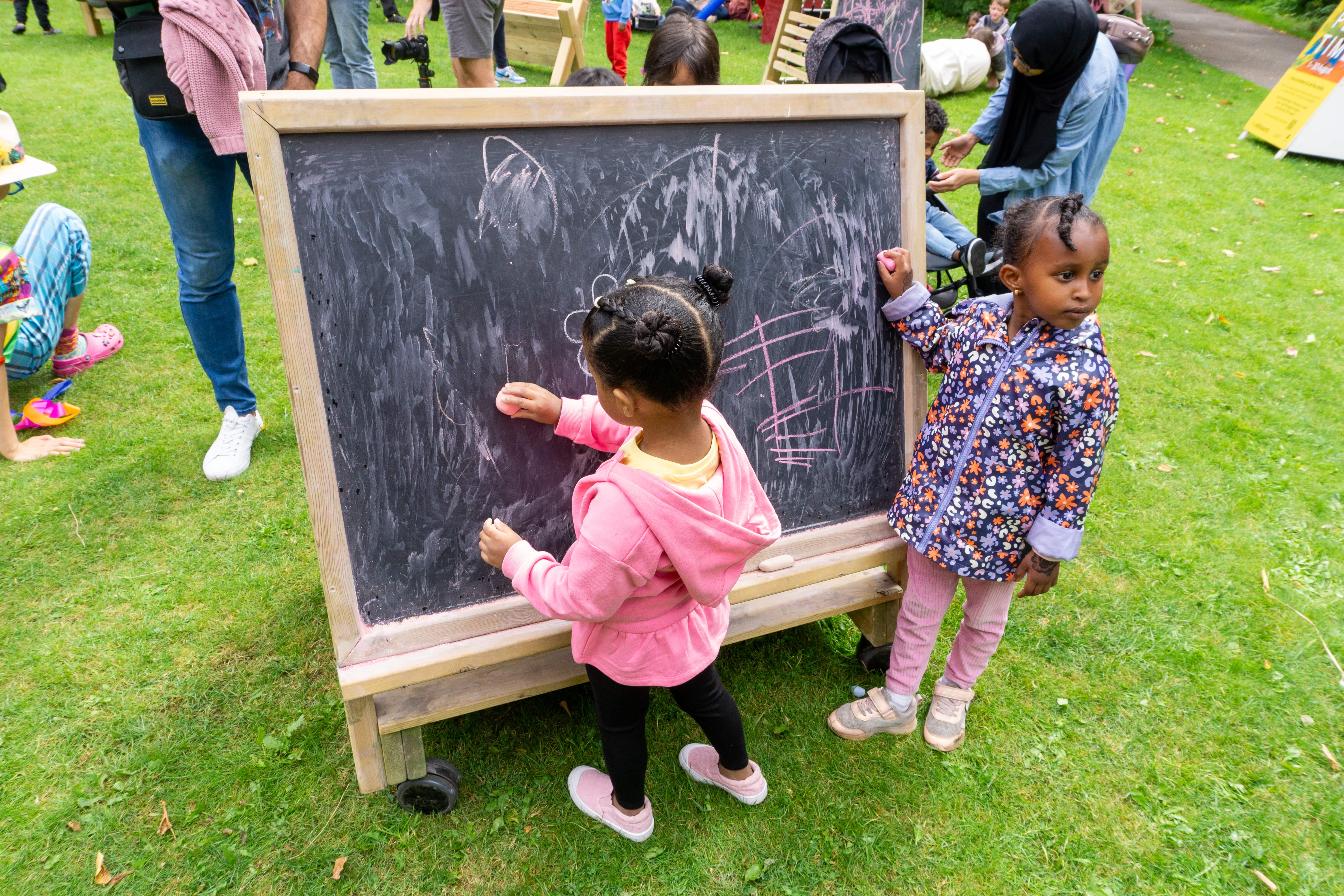 Two little children are drawings on a chalkboard that is on wheels. The children are using pink chalk to draw a variety of shapes and patterns. The chalkboard is on artificial grass