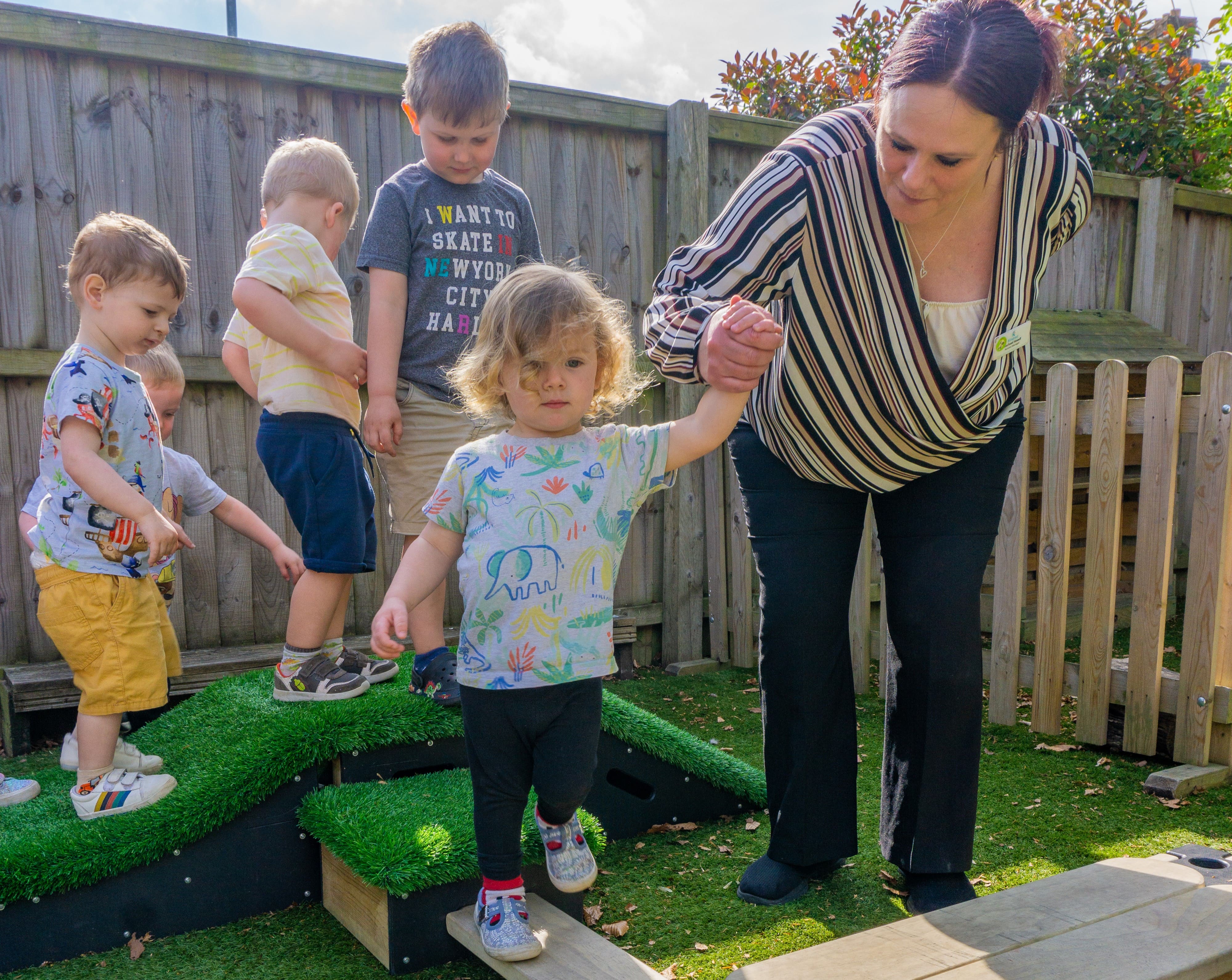 A little girl is being helped across a play builder plank, which is connected between two Get Set, Go! Blocks. The teacher is talking to the child and making them smile, whilst a line of 4 children wait for their turn.