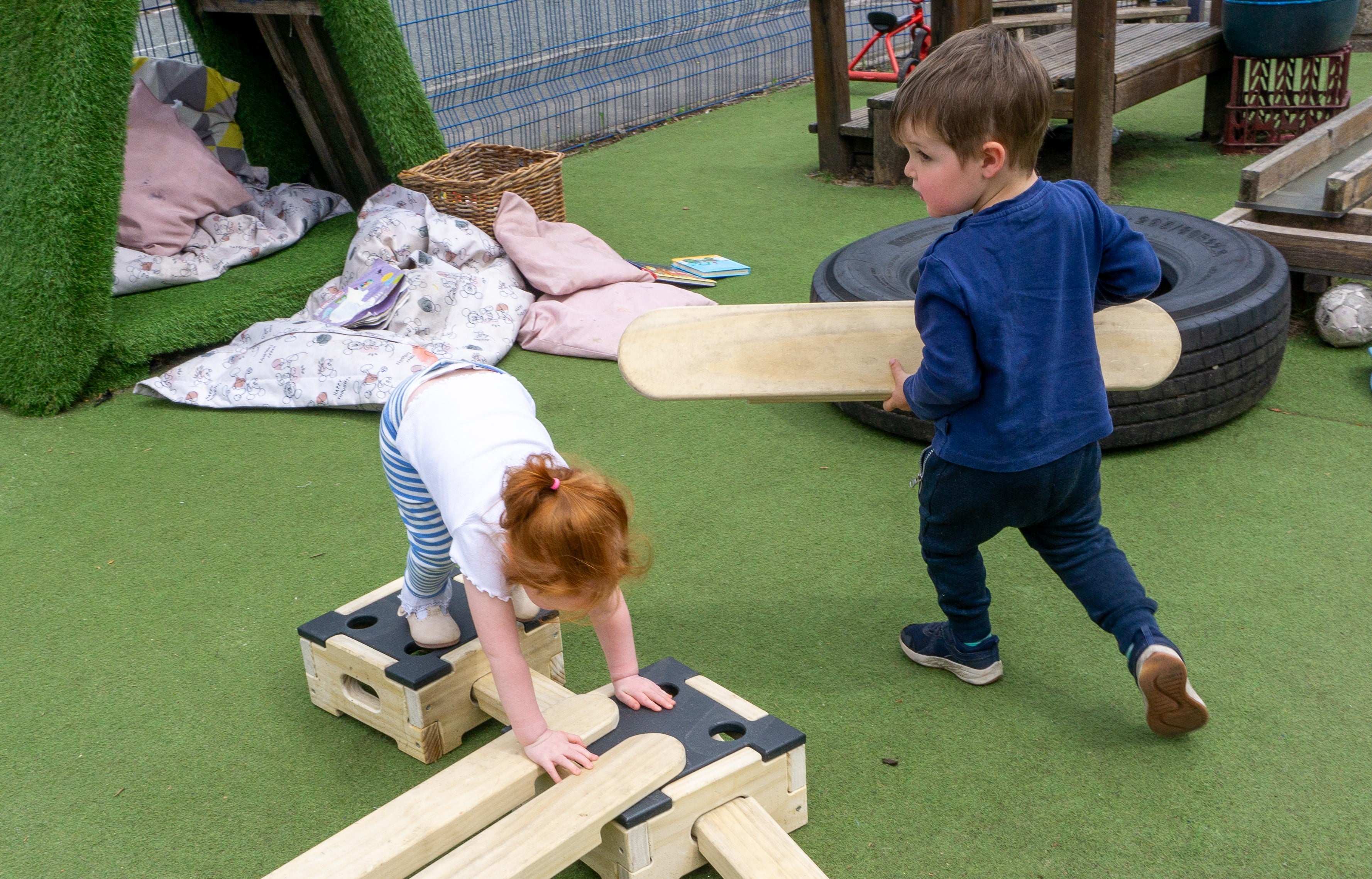 A child is connecting wooden planks to wooden blocks as another child is holding a wooden plank and is running around the other child. This is taking place on an artificial grass surface.