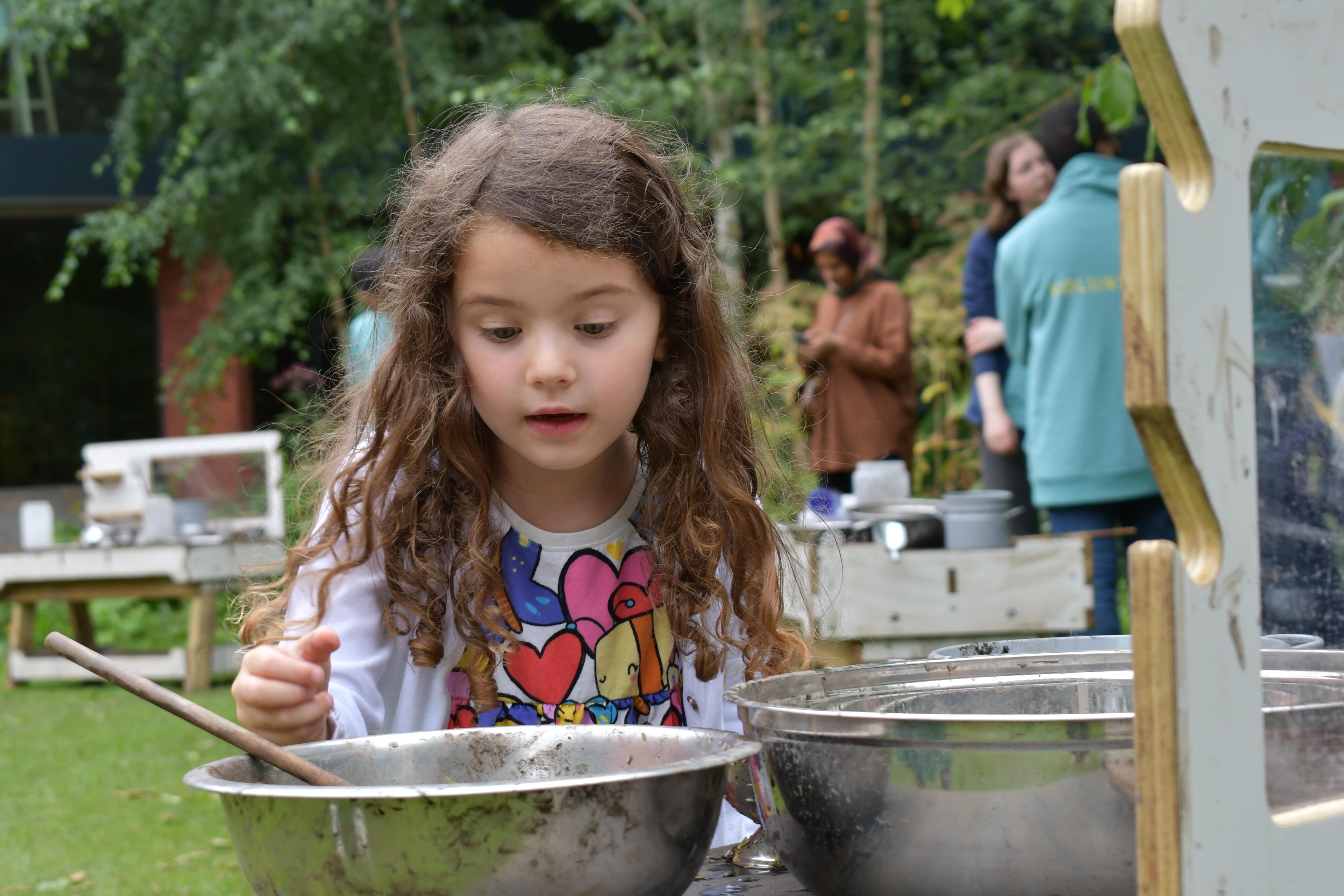 A girl is playing with two metal bowls that have been placed on a mud kitchen. A wooden spoon has been placed in the bowl and people can be seen in the background/