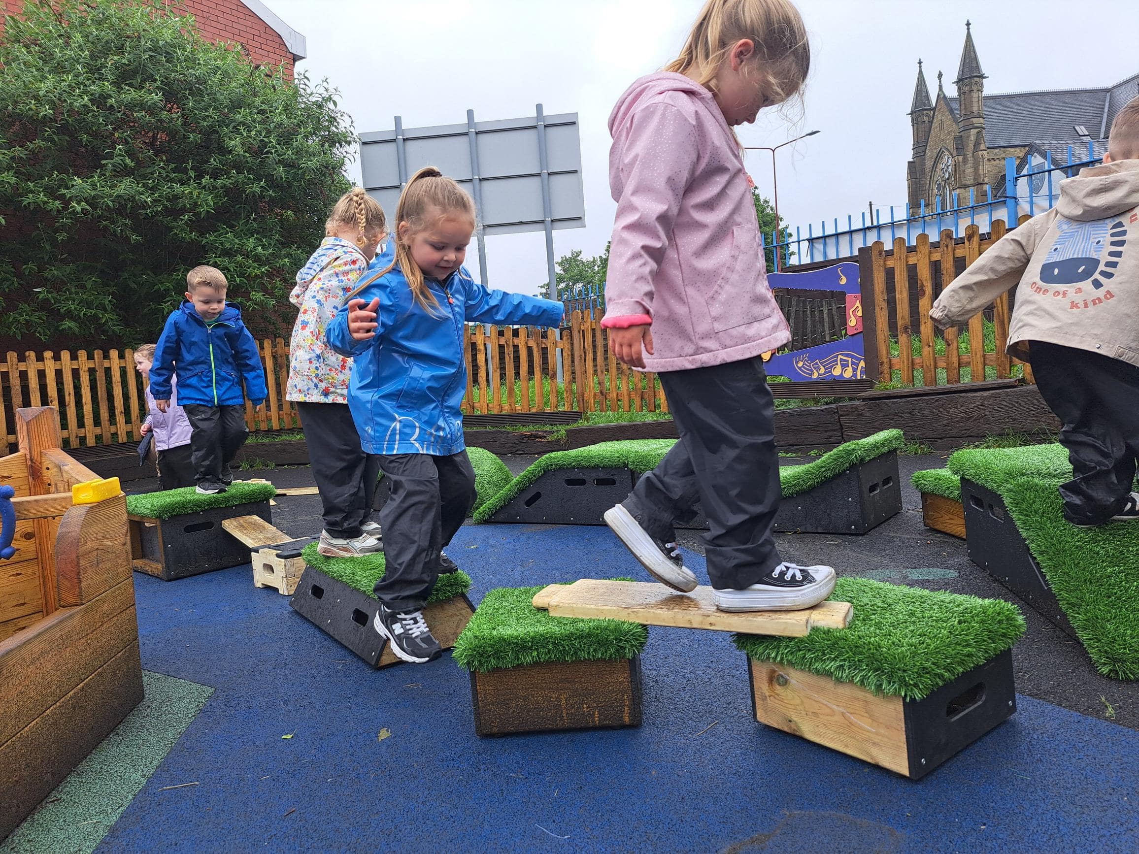 A group of children are playing on wooden planks and blocks. The children are smiling and cheering as they cross the obstacle course that they created.