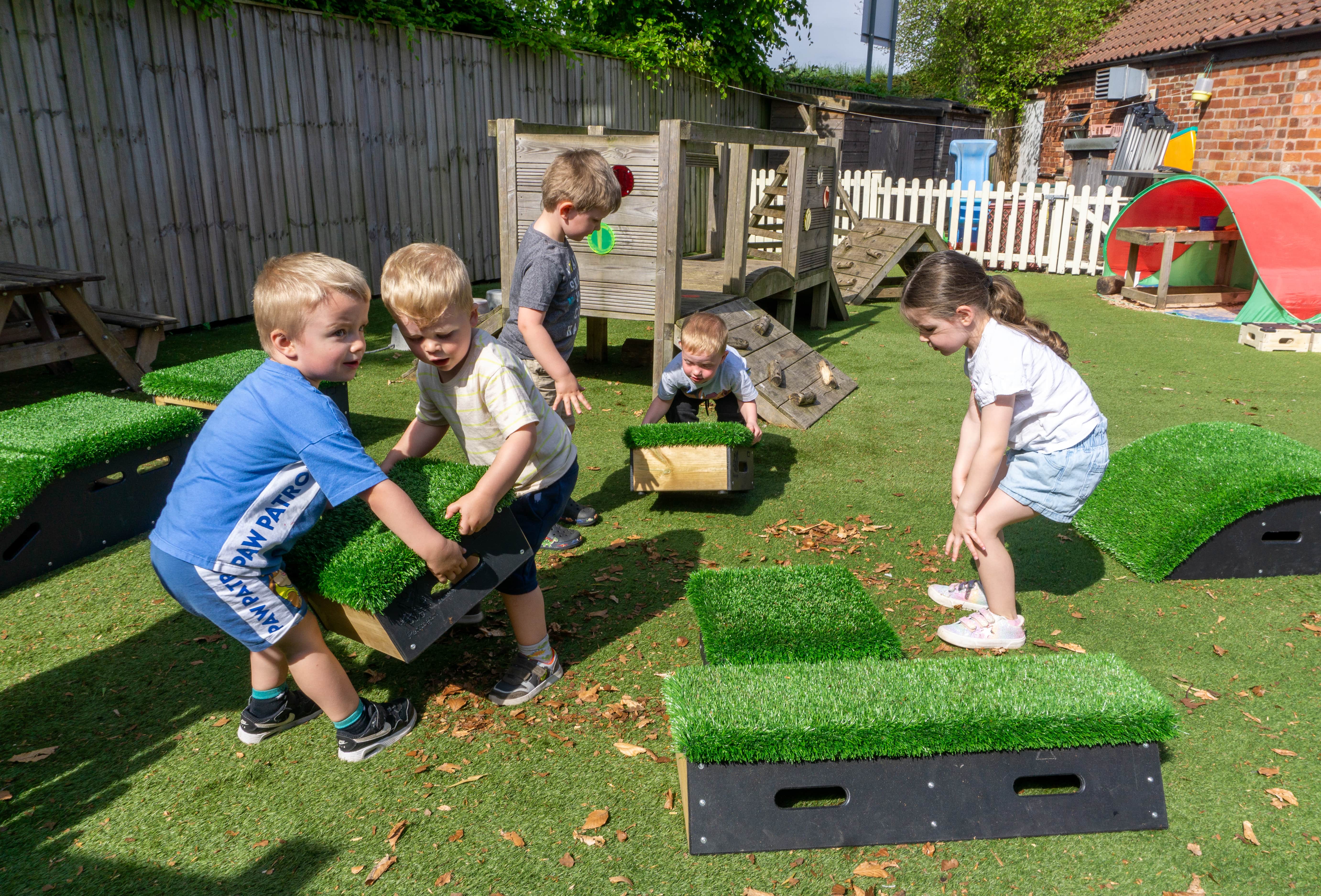 Two children are lifting a wooden block with artificial grass placed on the top. A little girl is giving the two children directions, whilst two other children start lifting another block.