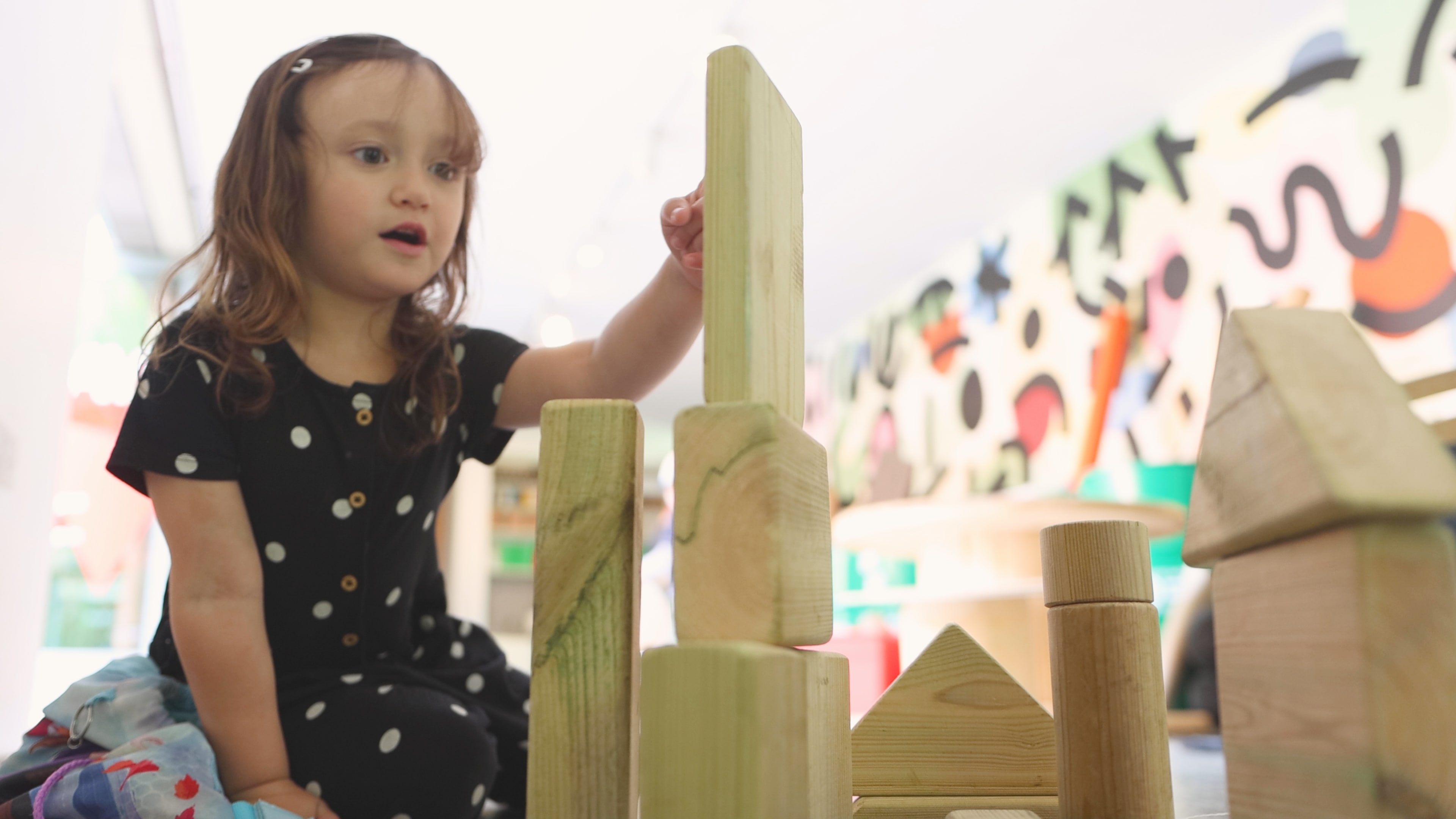 A little girl is playing with wooden STEM blocks. She is building a tower out of the blocks inside of the classroom.