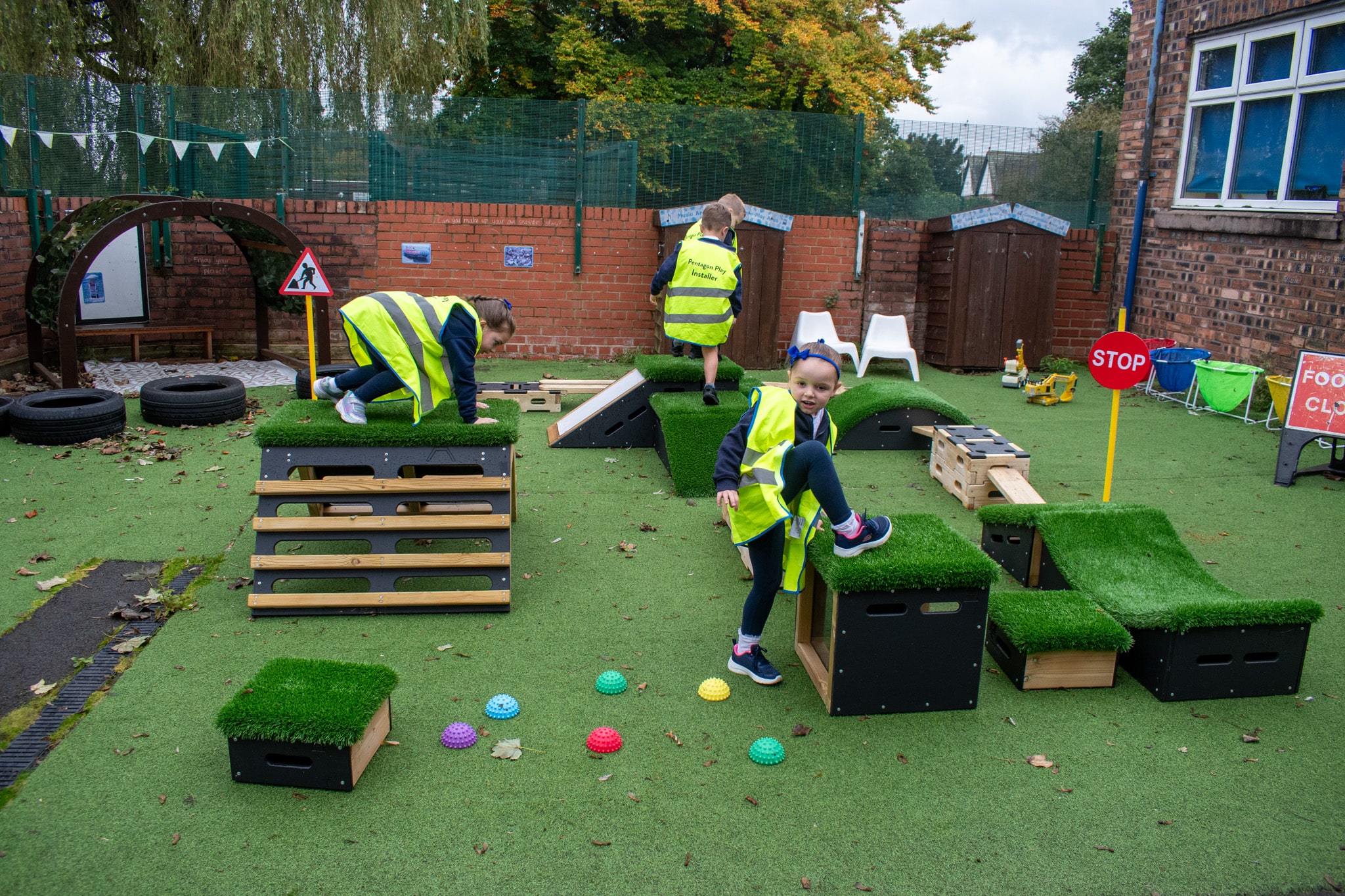 A group of children are wearing hi-vis jackets as they explore all of the new freestanding active play equipment. The children are smiling and cheering.