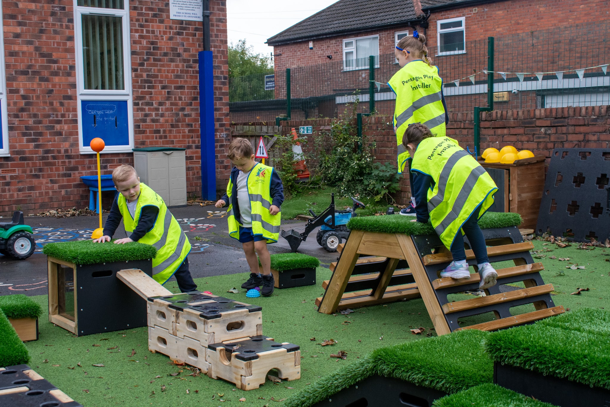 4 children are all wearing hi-vis jackets as they all climb on different pieces of play equipment. The equipment is placed on top of an artificial grass surface.