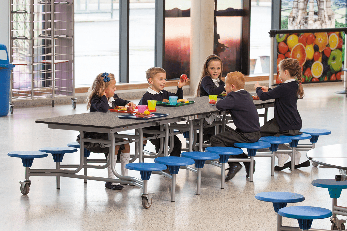 A group of children are sat on a 16 seater school dining table and are eating lunch! The children are talking to each other.