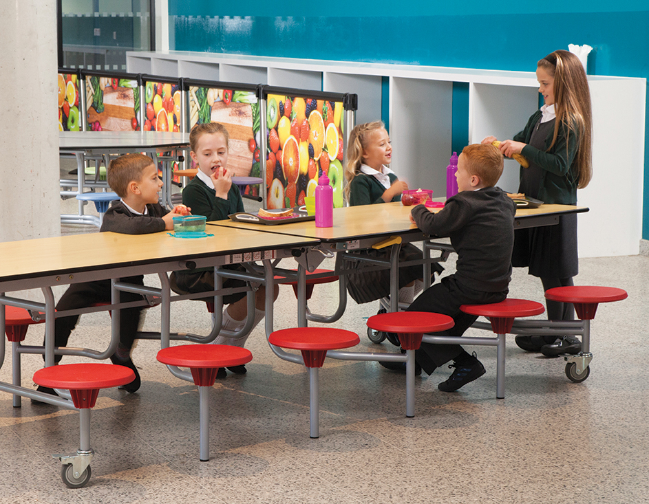 A group of children are sat around the school dining table and are talking. The stools are red and the table top is maple.