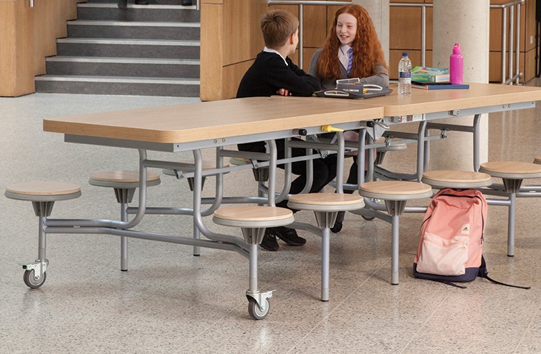 2 children are sat at a school dining table and are talking to each other. The table is maple.