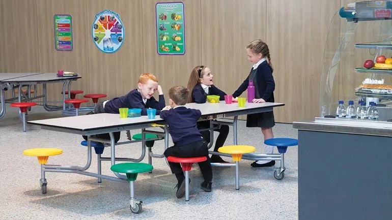 4 children are all sat around a school dining table and are talking. The table is rectangular with different coloured stools.