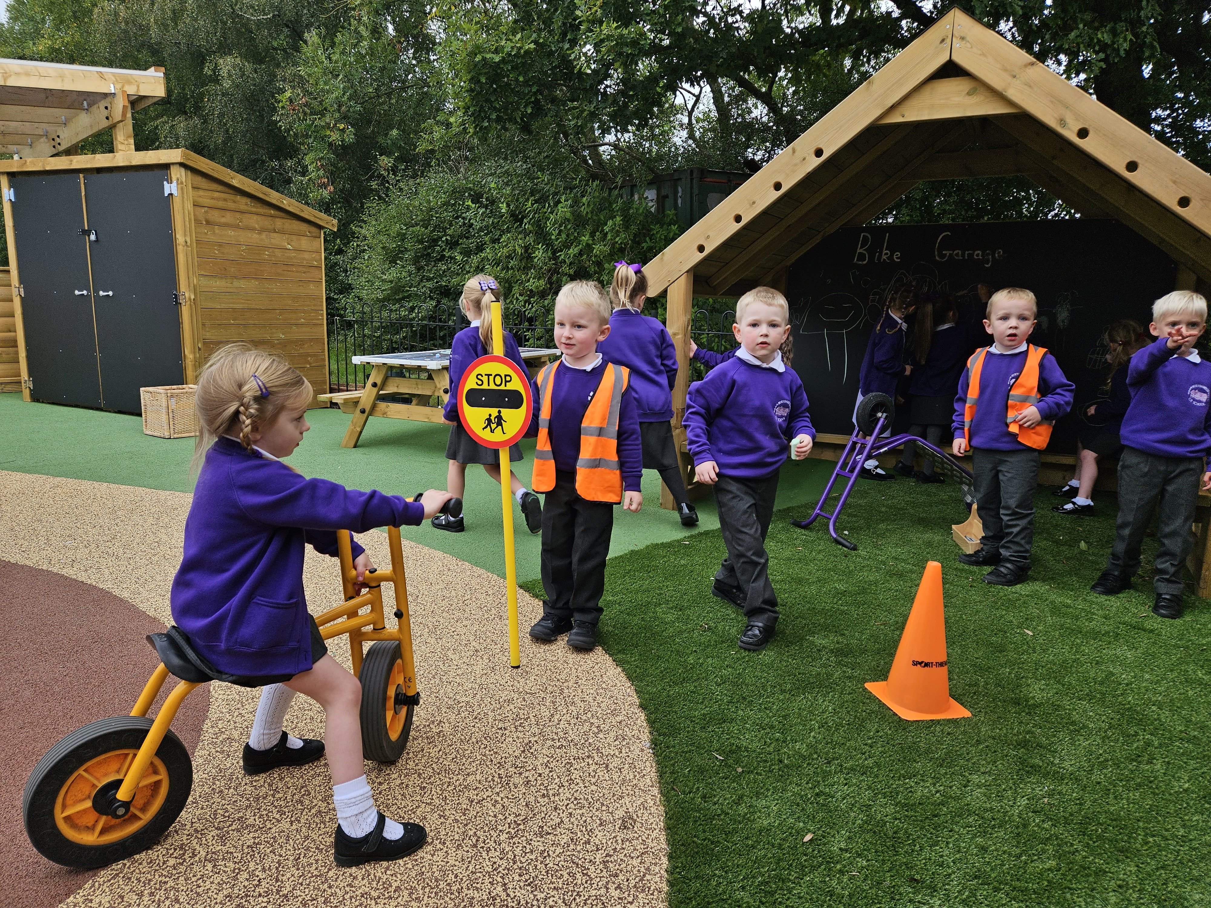 A group of children are stood on artificial grass and are waiting beside a wetpour road. A child is riding a bike on the wetpour road and is being stopped by another child who is dressed up as a lollipop man.