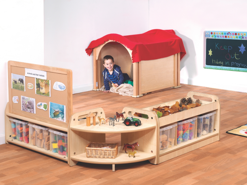 A child is hiding within a wooden cave-like structure, with bookshelves and storage units placed around the cave. All of the furniture has been built from wooden materials.