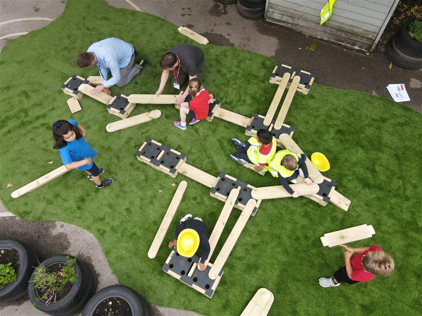 Group of children playing on a Play Builder Architect set, which has been set up on artificial grass. The children have been joined by a teacher and a member of the Pentagon Play team.