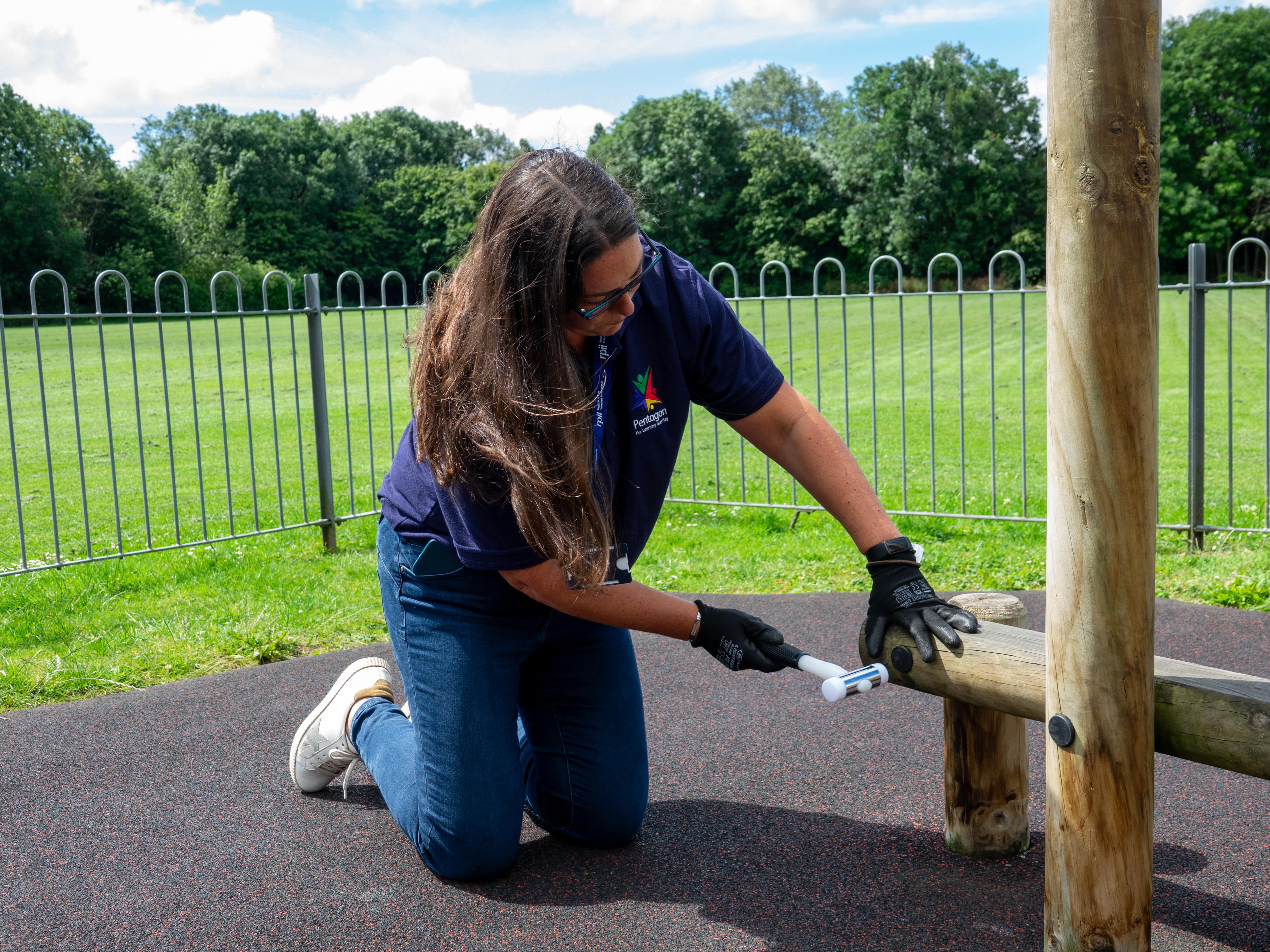 A playground inspector is hammering in a loose fitting for a piece of play equipment.