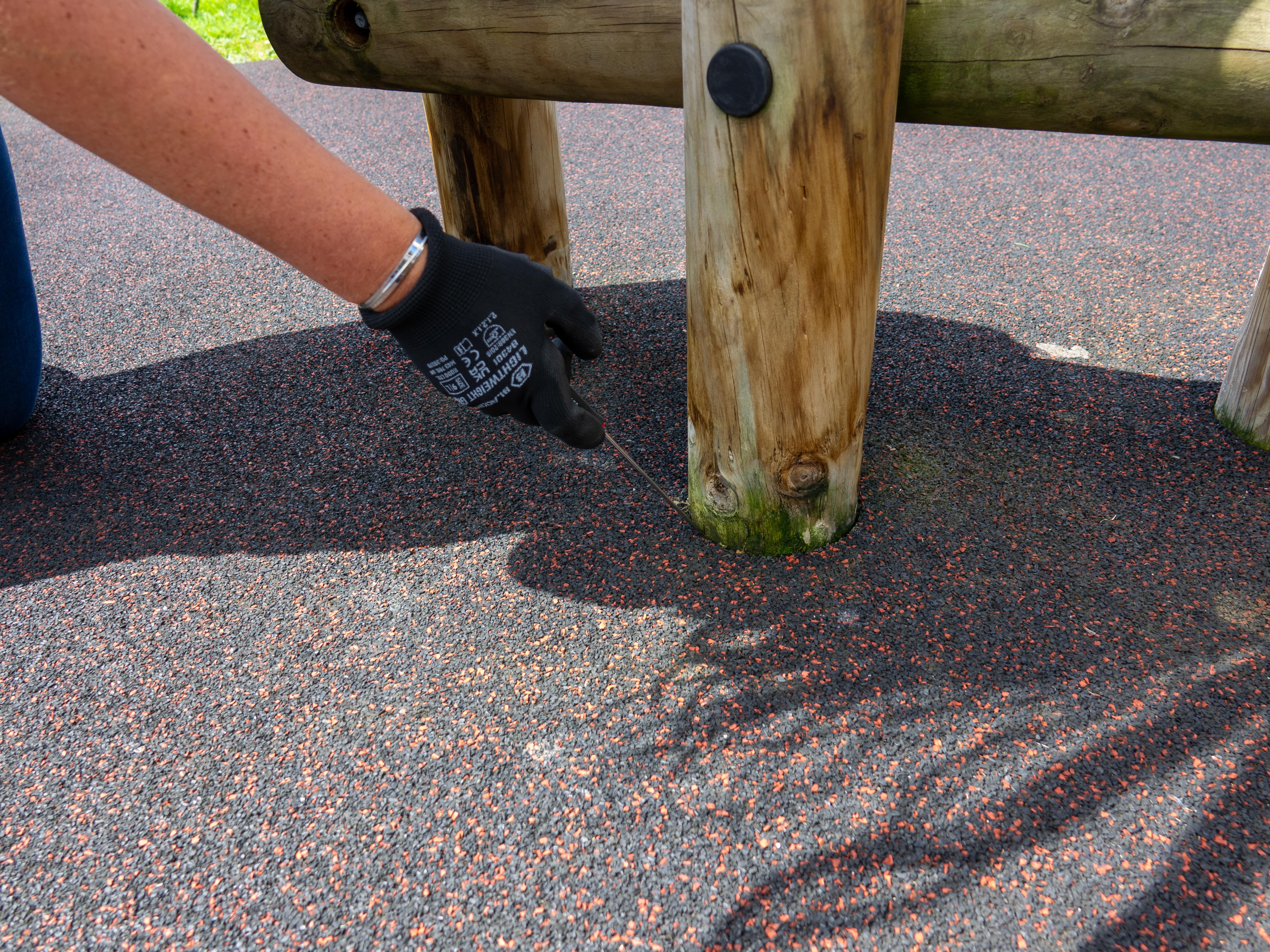 A playground inspector is pointing out mould that has grown on the bottom of a piece of play equipment. The play inspector is using a screwdriver to move the wetpour to reveal the damage.