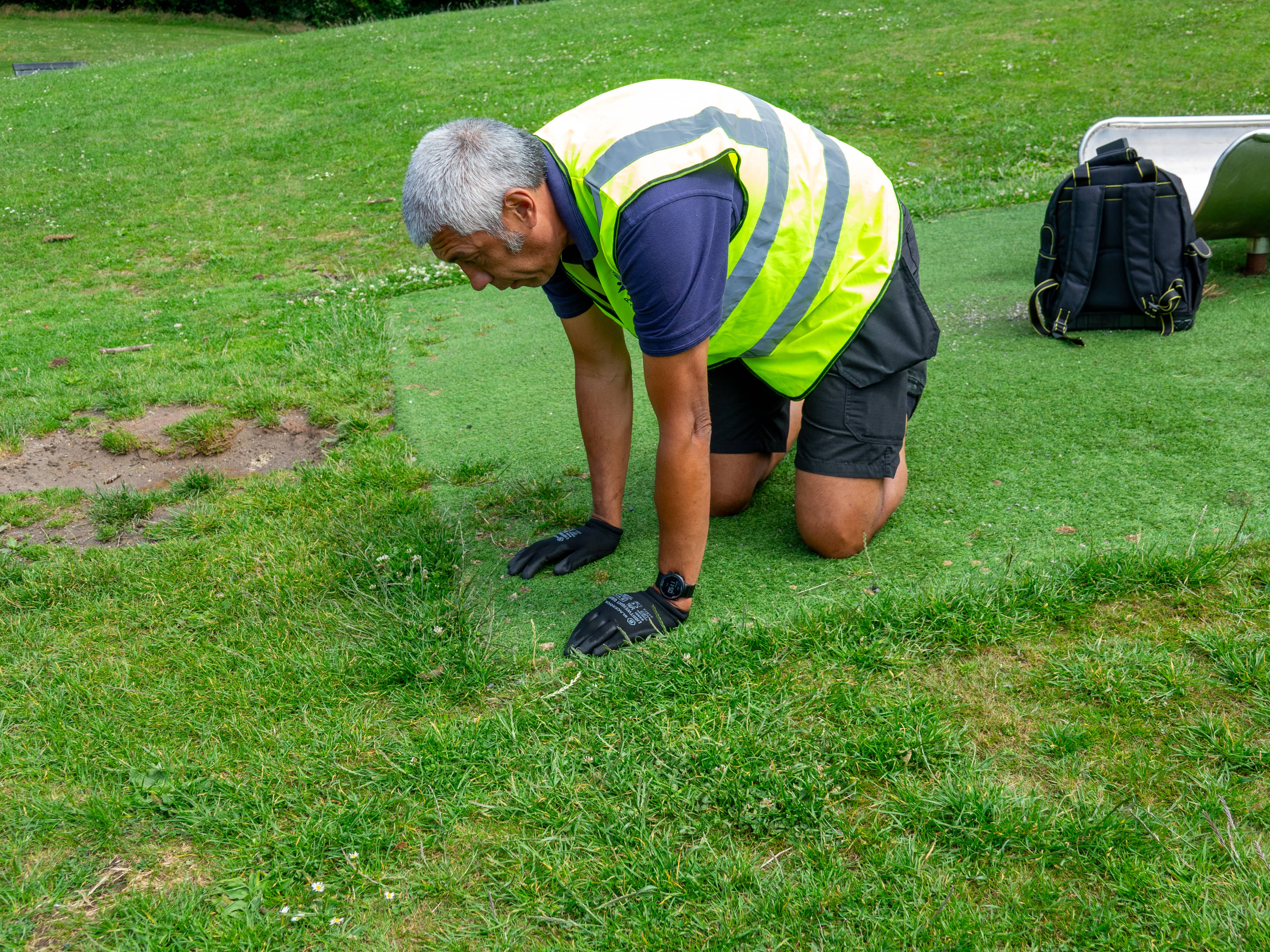 A male playground inspector is inspecting the artificial grass surfacing to look at the draining system.