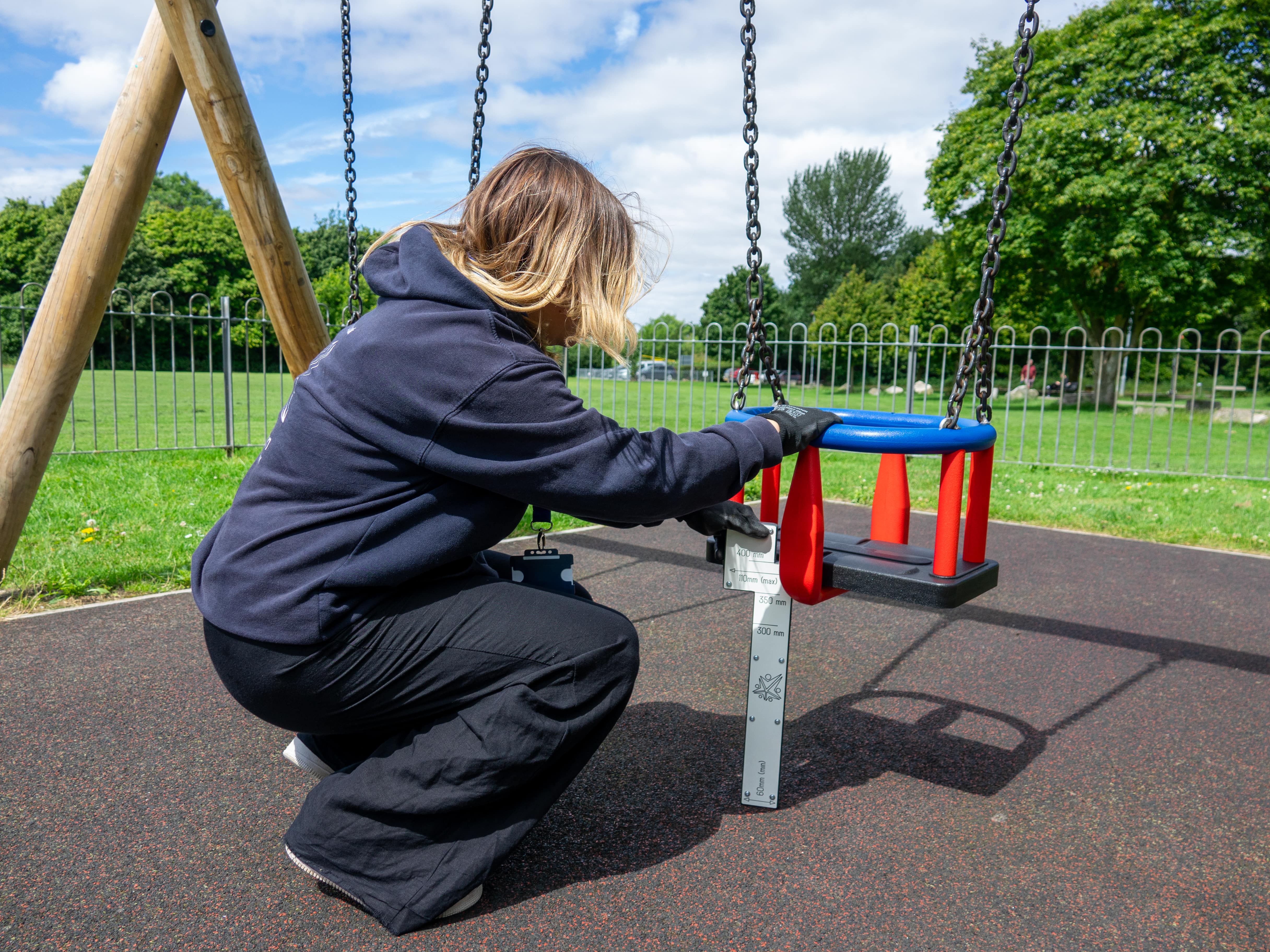 A female playground inspector is measuring the distance between a swing seat and the safety surface.