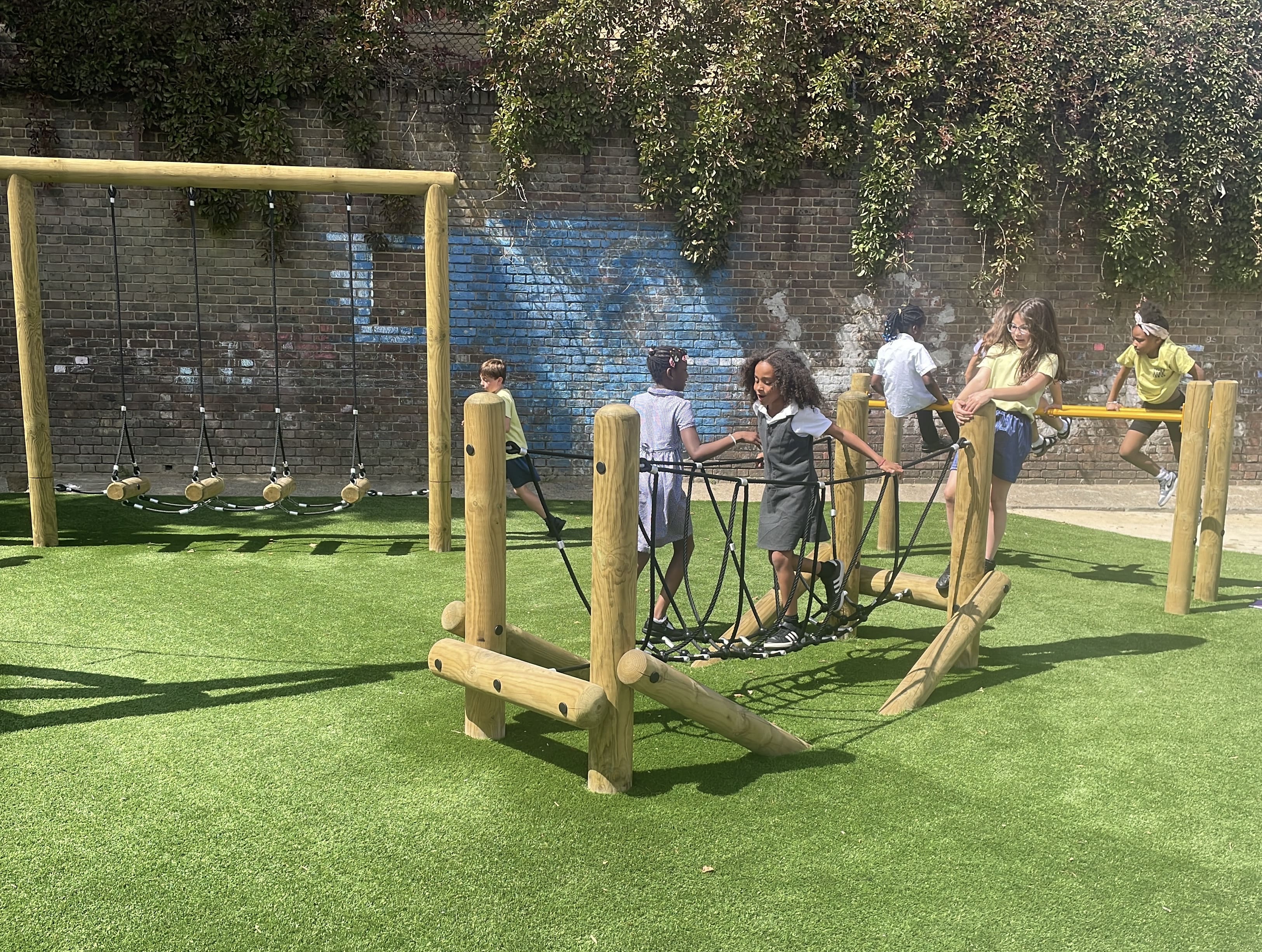 Children are playing on a variety of different pieces of trim trail equipment. A couple of children are on the rope bridge, whilst a couple are on the parallel bars.