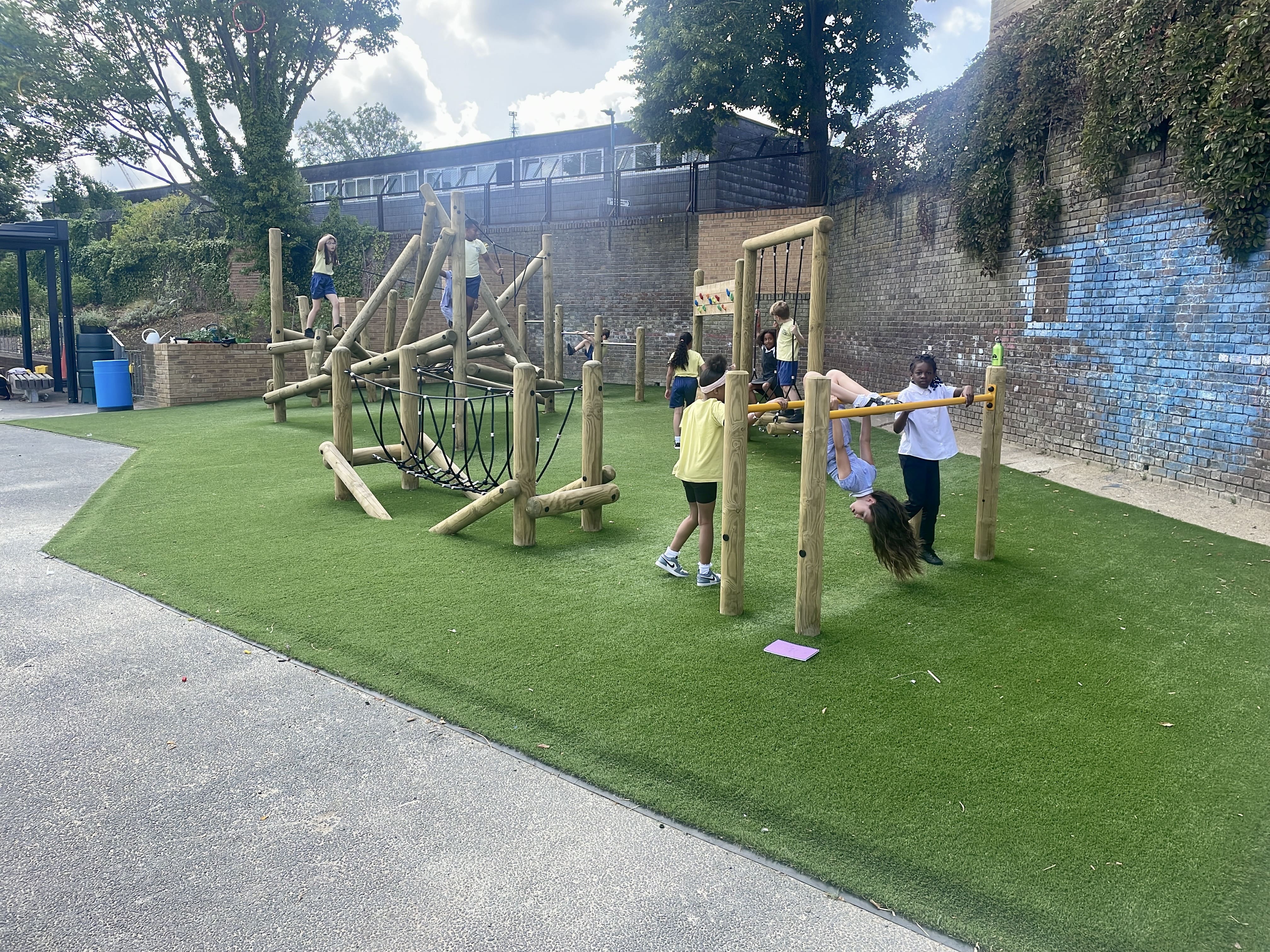 An artificial grass surface is beneath a variety of playground equipment. A group of children are currently playing on top of it.