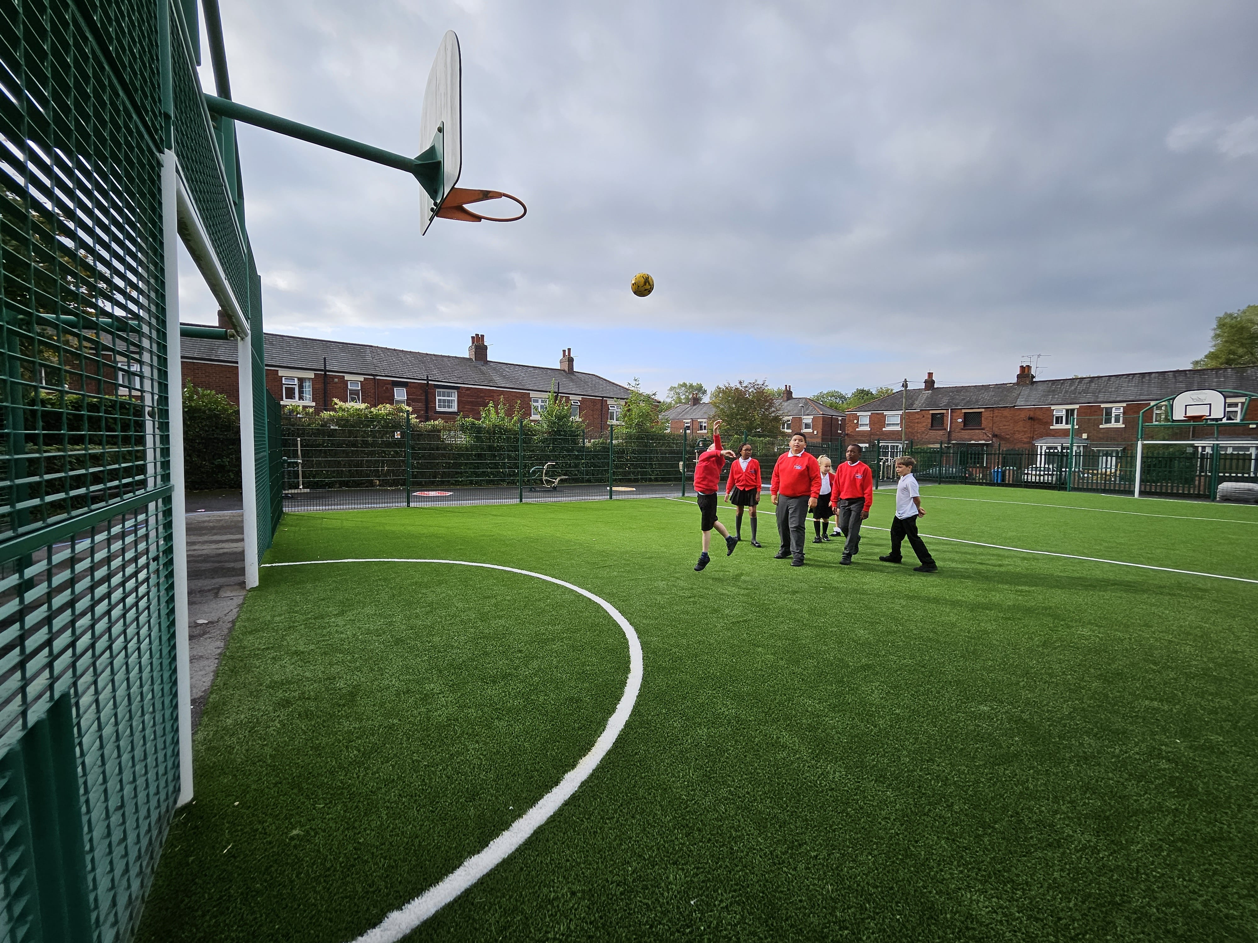 A group of children are playing basketball on a MUGA pitch. One child has thrown the ball and the ball is heading towards a basketball hoop.