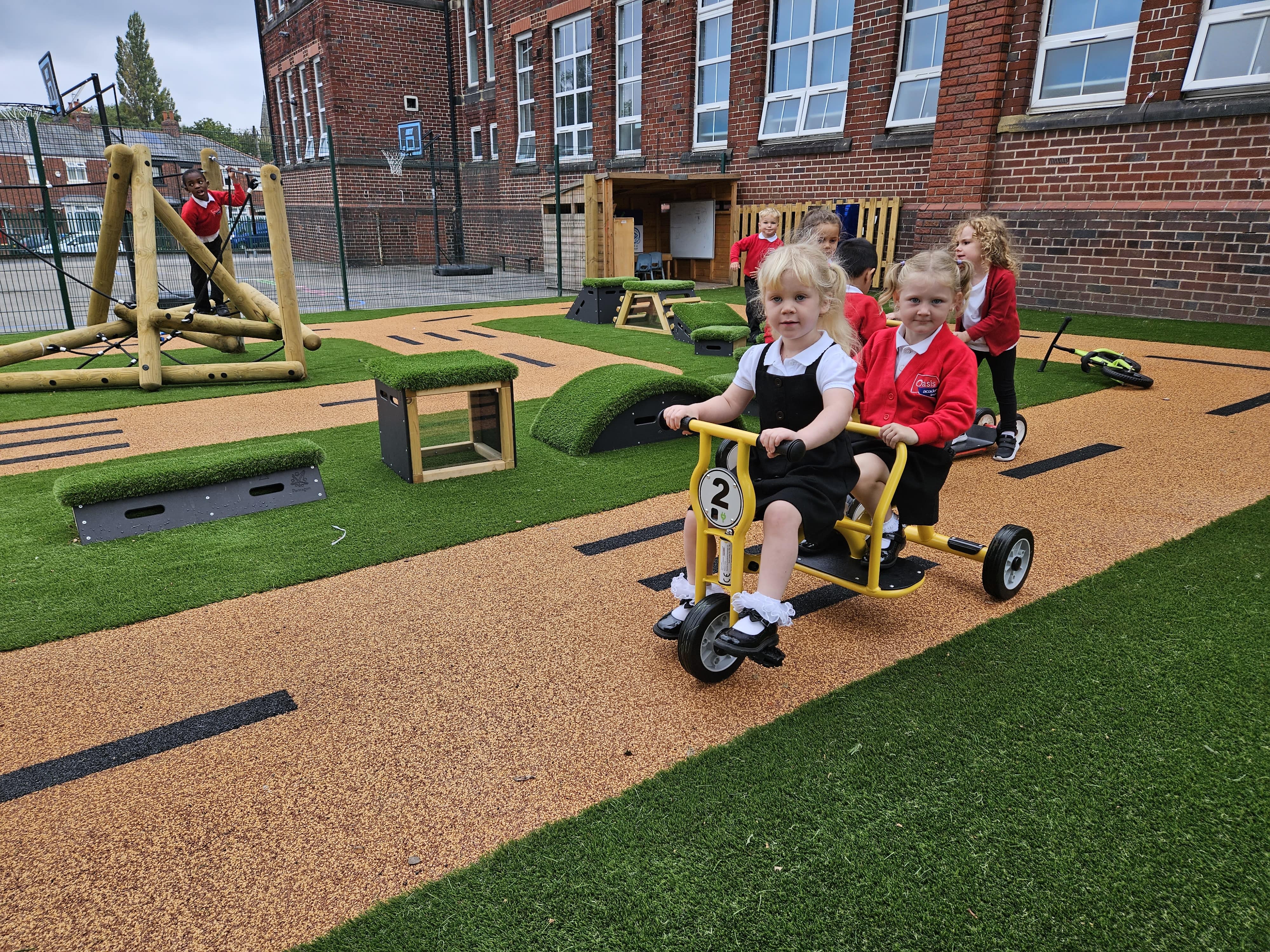 2 children are riding a tricycle on a wetpour road, with a group of children in the background playing on a variety of freestanding play equipment. Some children are playing on a climbing frame.