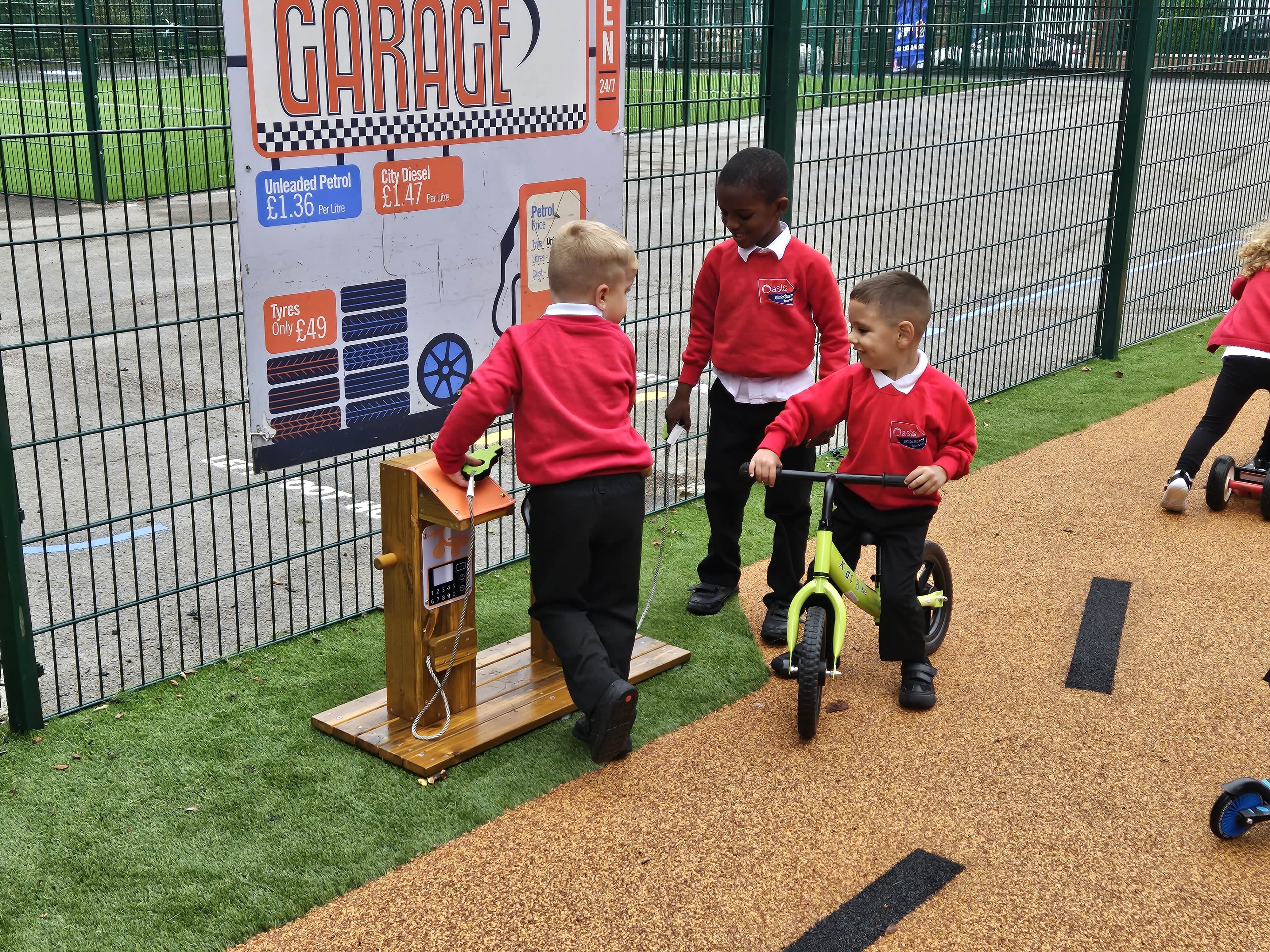 A group of children are playing on tricycles and with a pretend petrol pump. On the other side of the role play equipment, a green mesh fencing is surrounding the playground.