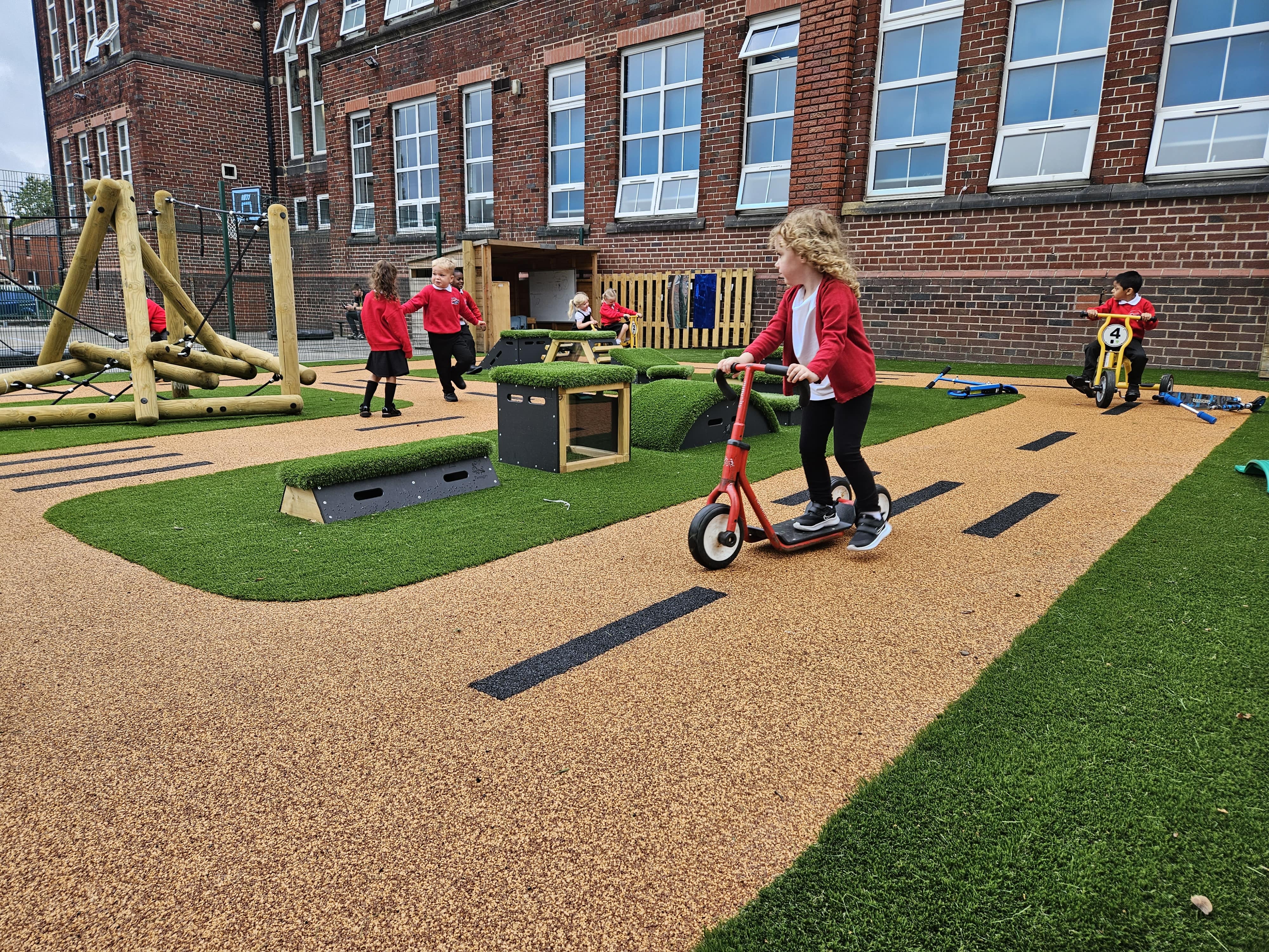 A little girl is riding a scooter on a wetpour road design, with other children playing around her on freestanding play equipment. One child is on a tricycle and is following the little girl.