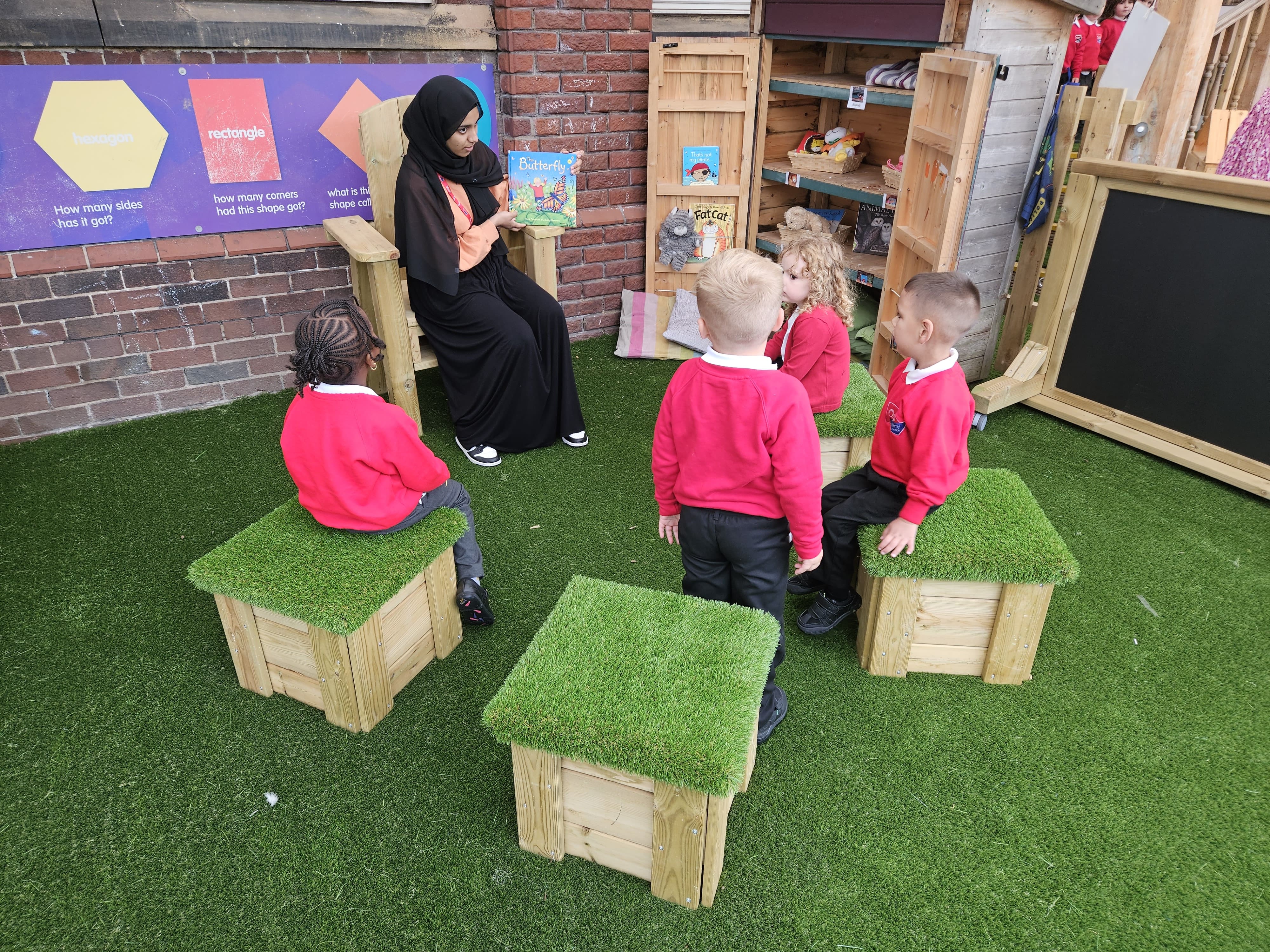 A teacher is sat on a Freestanding Storytelling Chair as she reads a book to a group of 4 children, who are each sat on an artificial grass block seat.