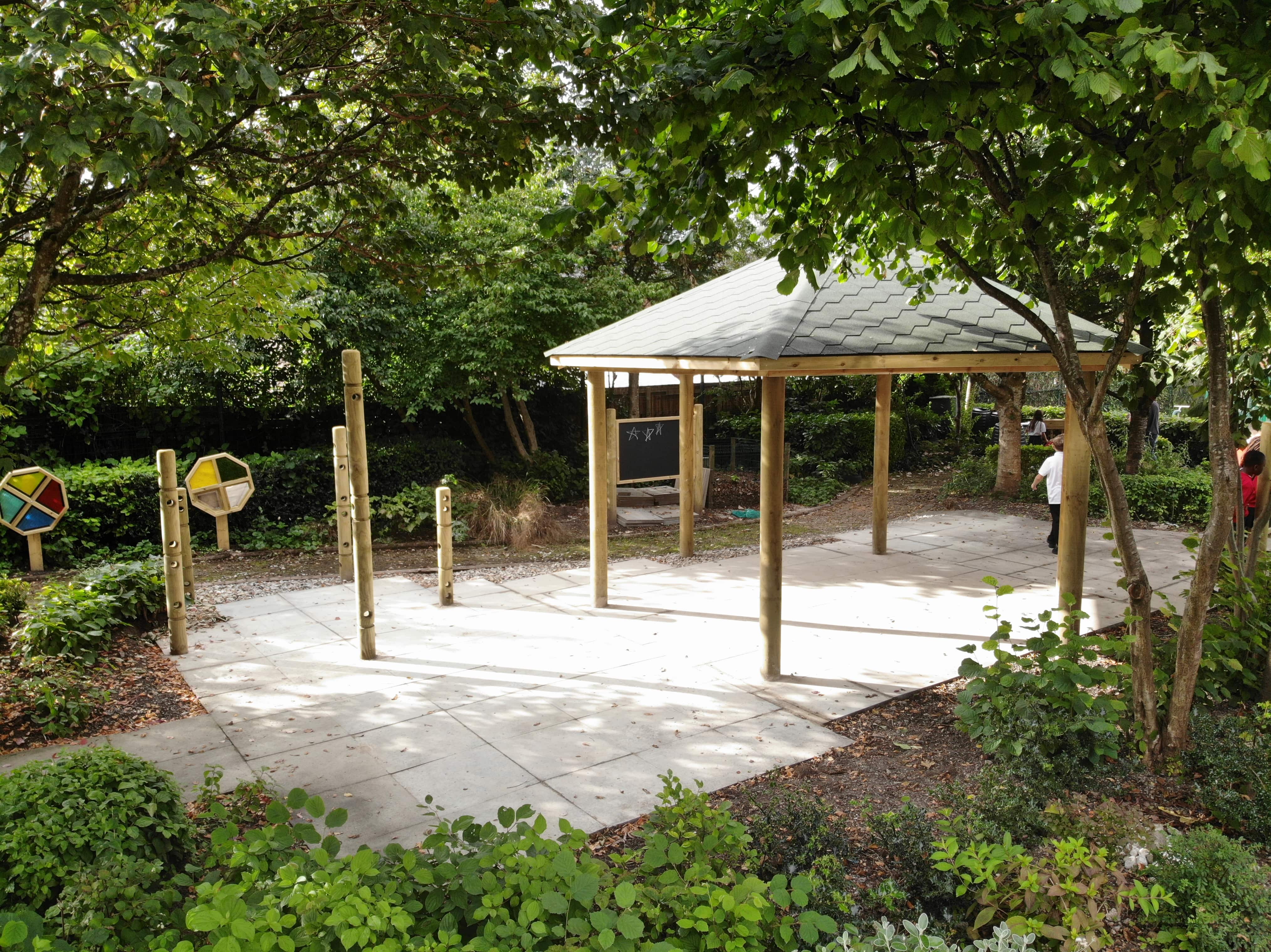 A wooden gazebo that has been installed in the middle of a forest. A variety of play equipment can be seen around it.