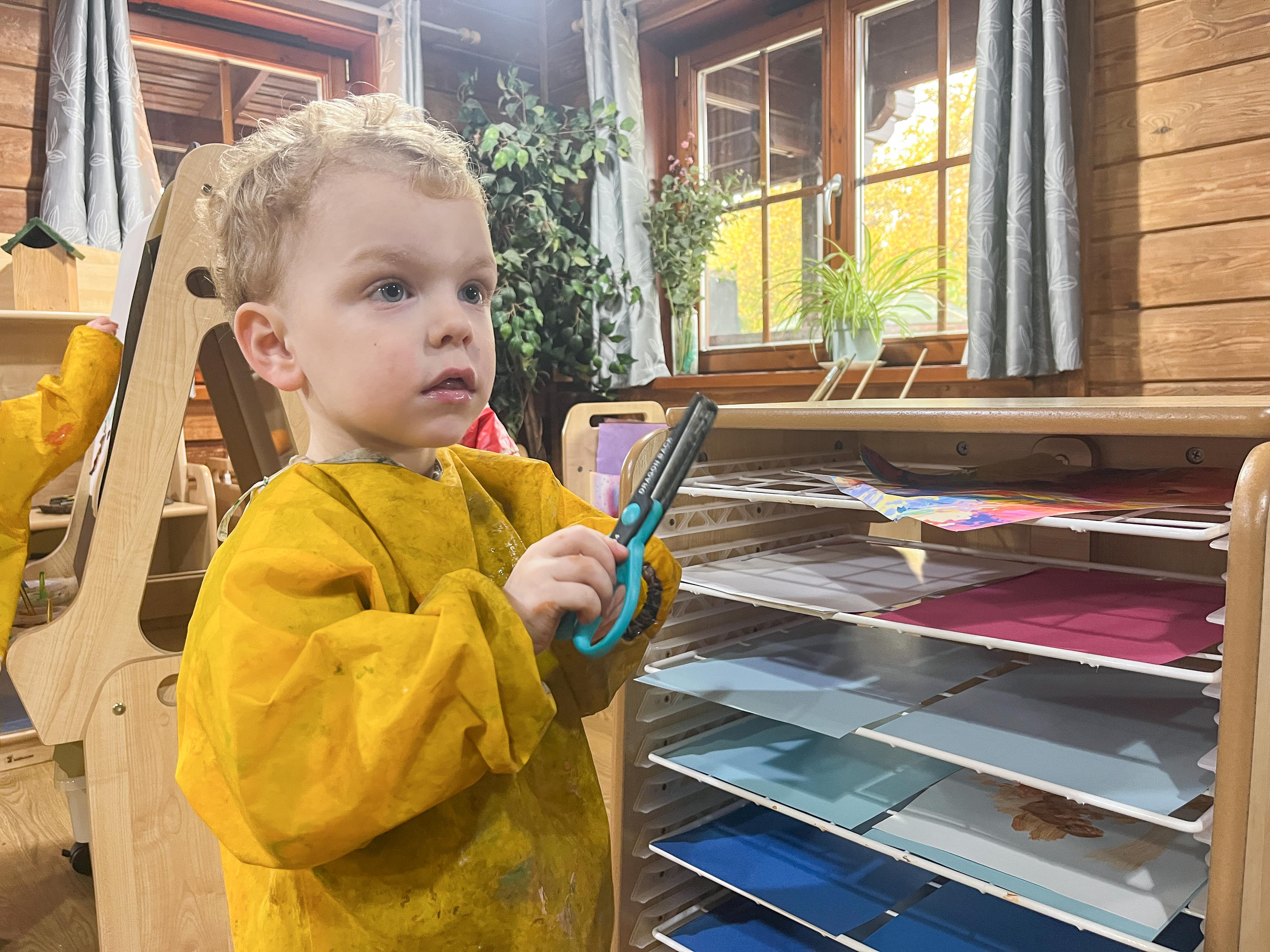 A little boy in an apron is holding a pair of scissors as he stands next to the Drying Rack. The boy looks amazed.