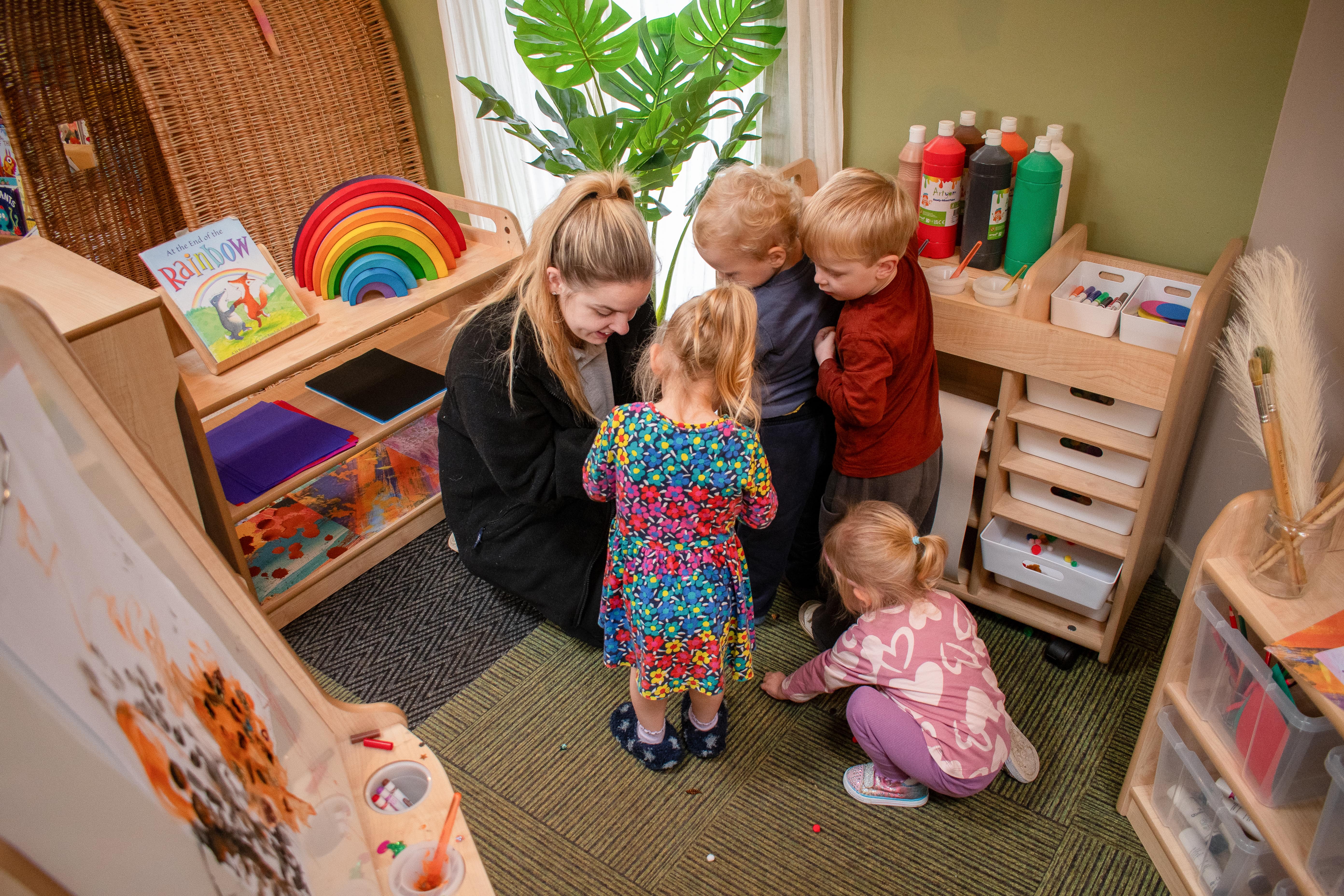 A member of nursery staff is talking to 4 eyfs children as they explore different pieces of nursery furniture.