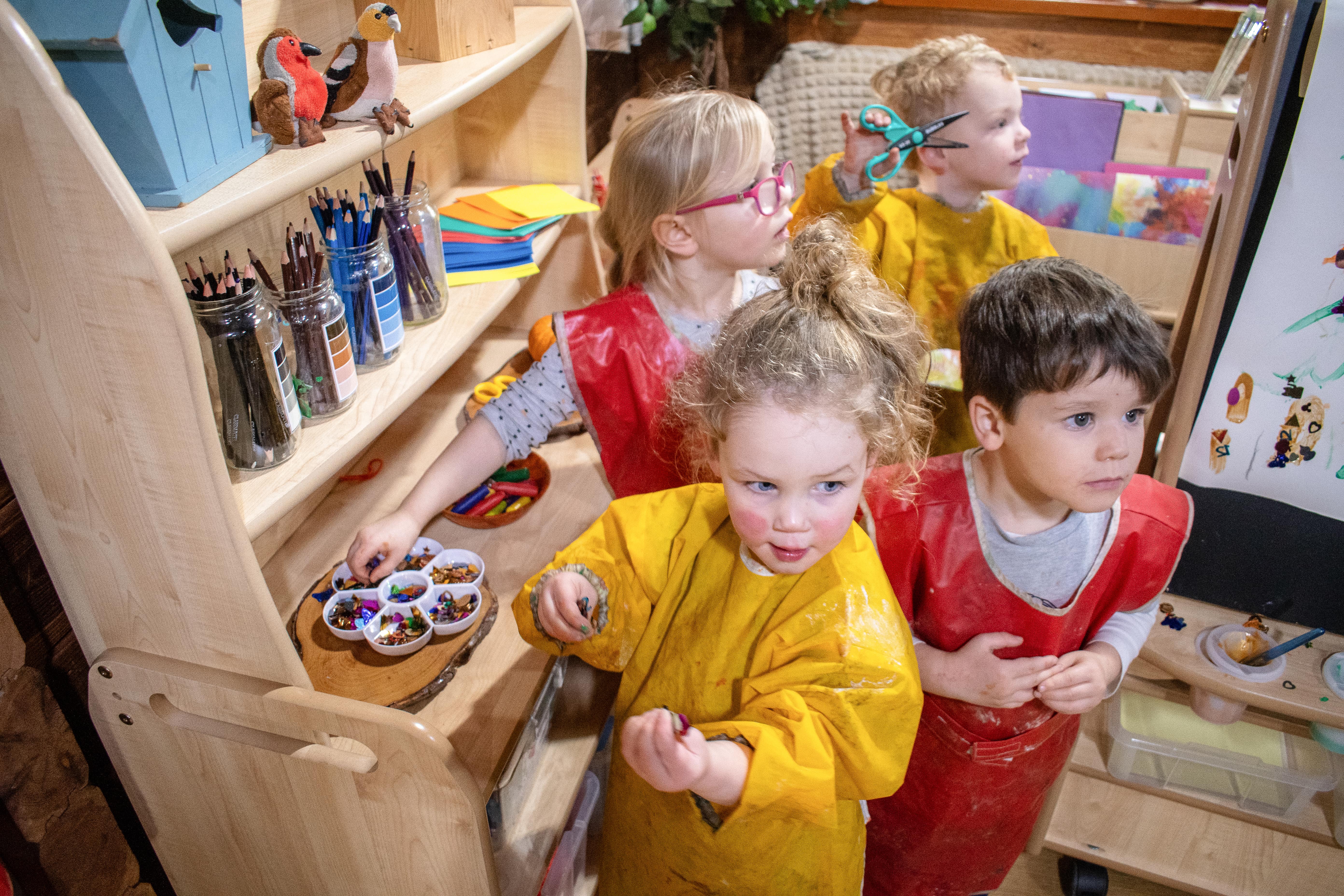 Four children are dressed in arts and crafts uniforms as they look off camera whilst holding a variety of art materials.