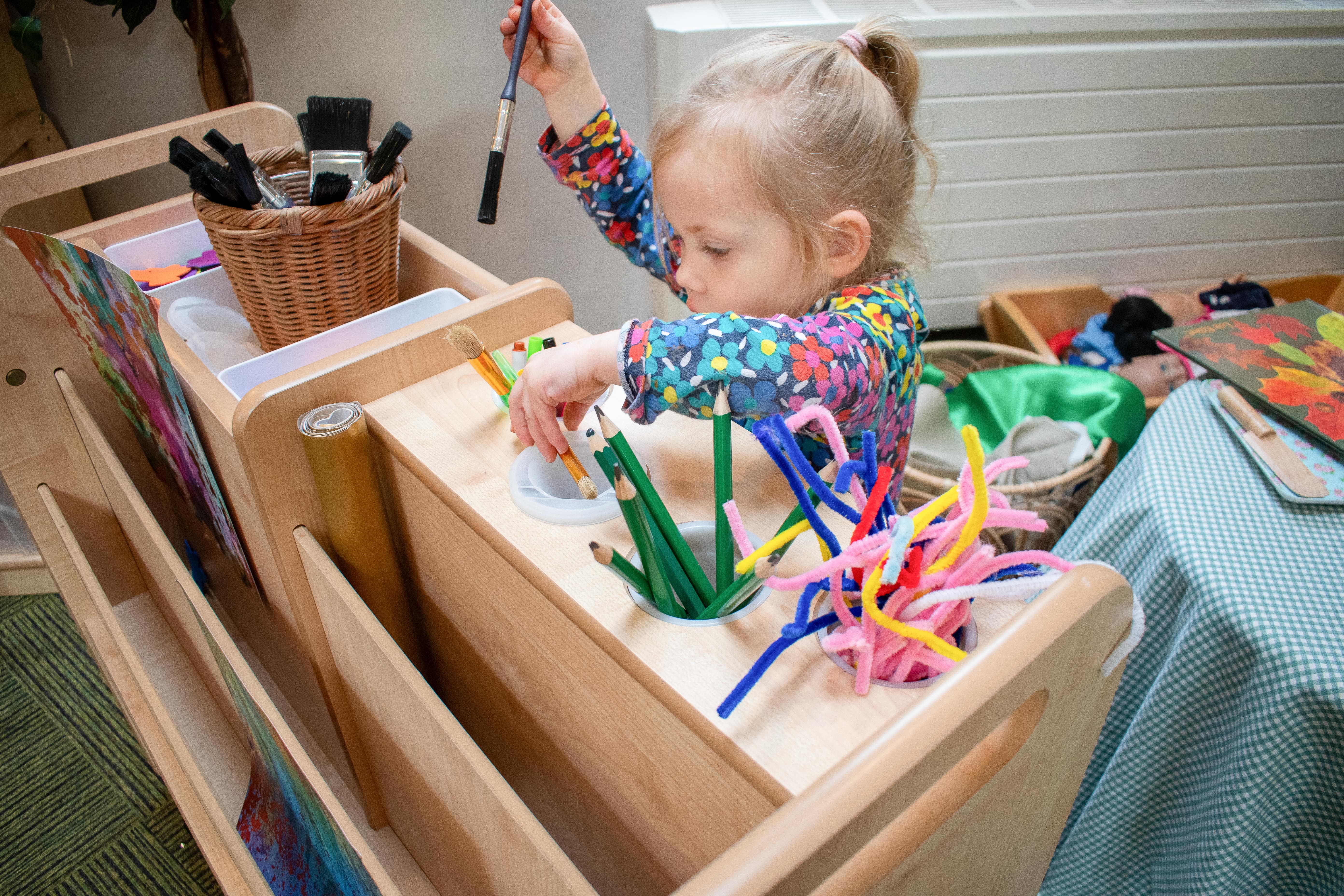 A little girl is retrieving art provisions from the Continuous Provisions Trolley, collecting random pencils and brushes.