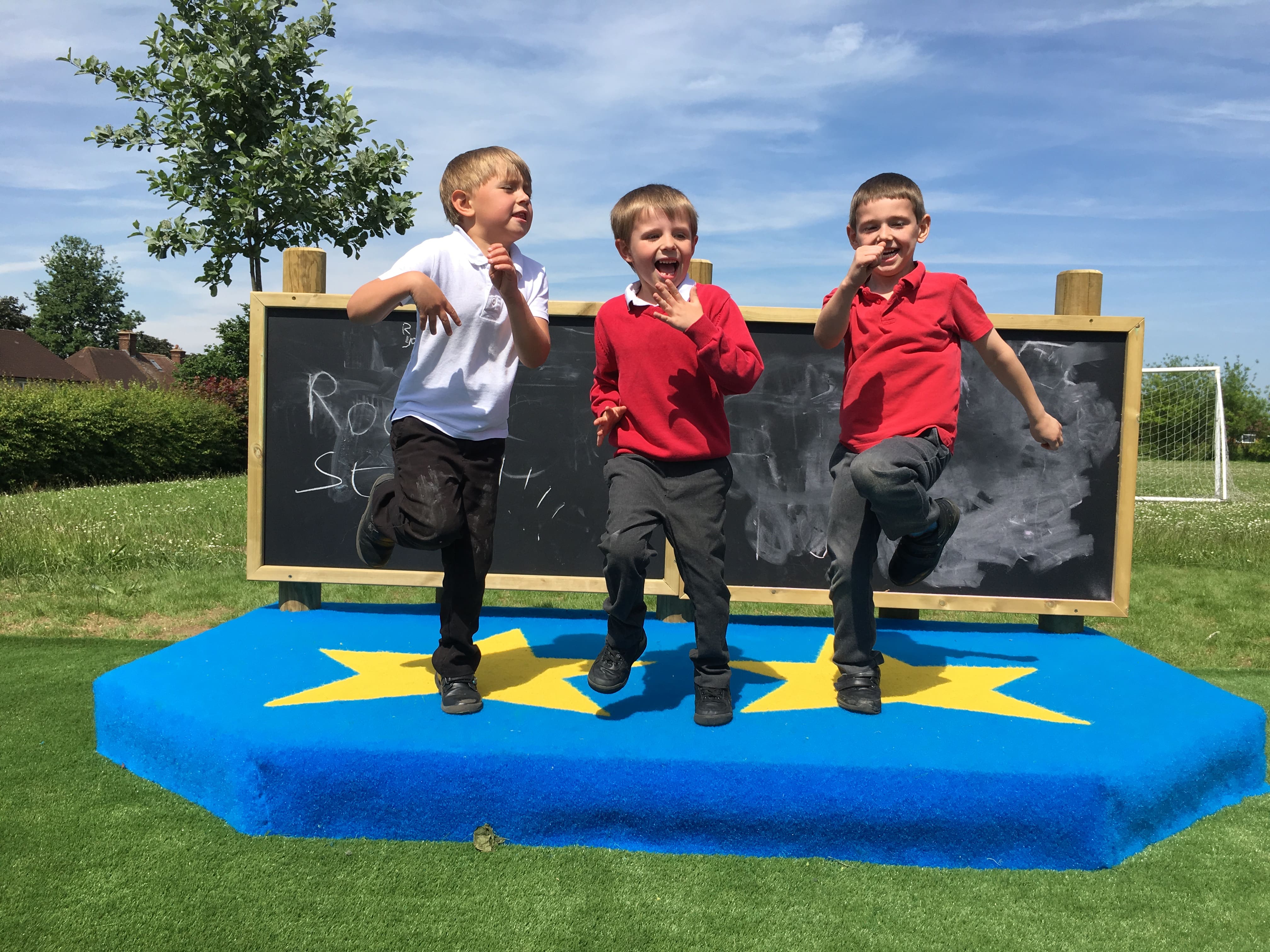 Three children are stood on a blue performance stage and are pointing at the camera and looking excited as they dance.