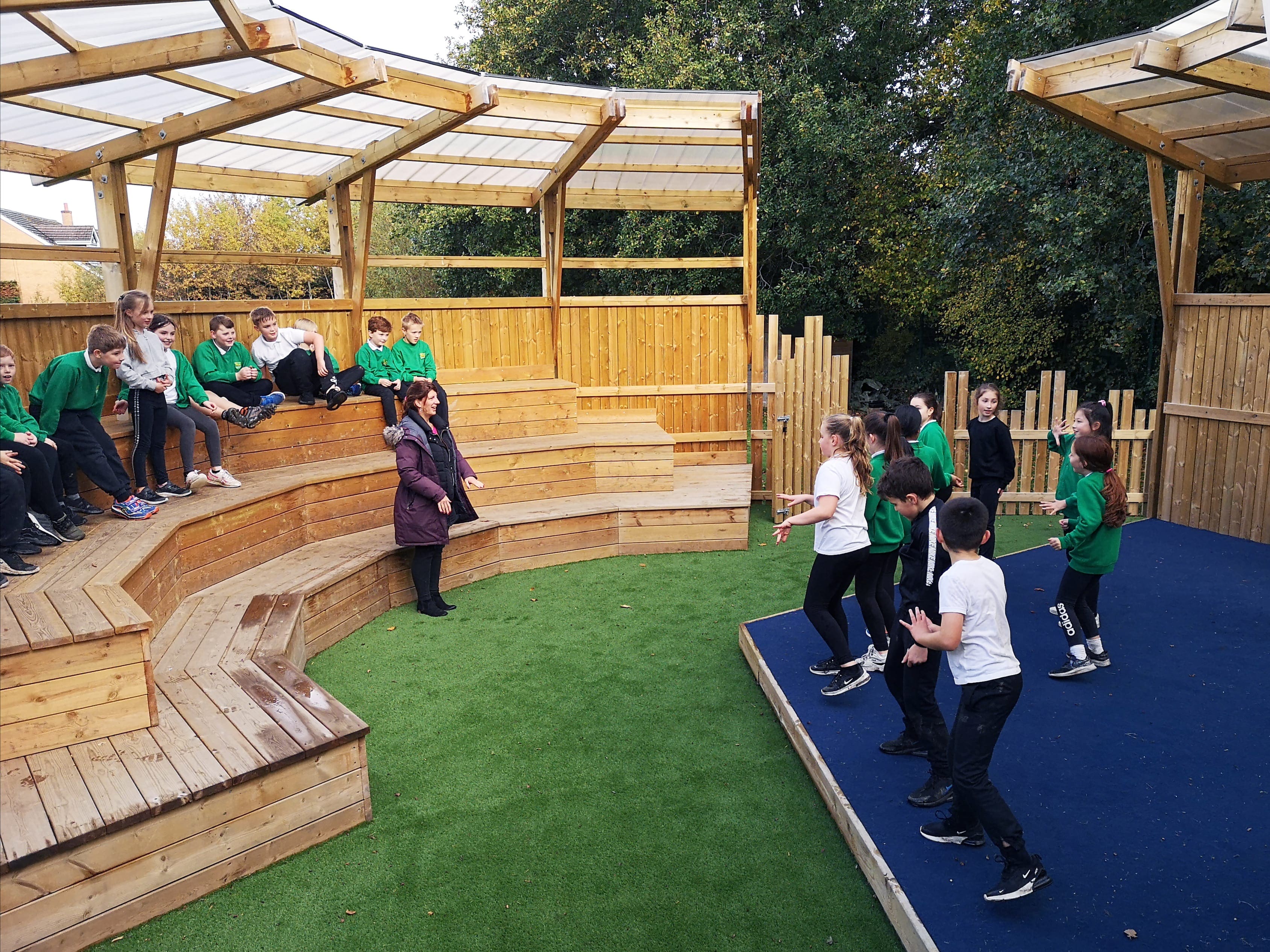 A group of children are stood on a performance stage and are rehearsing a dance. A teacher is stood at the front and guiding them, with a group of pupils sat on benches behind them.