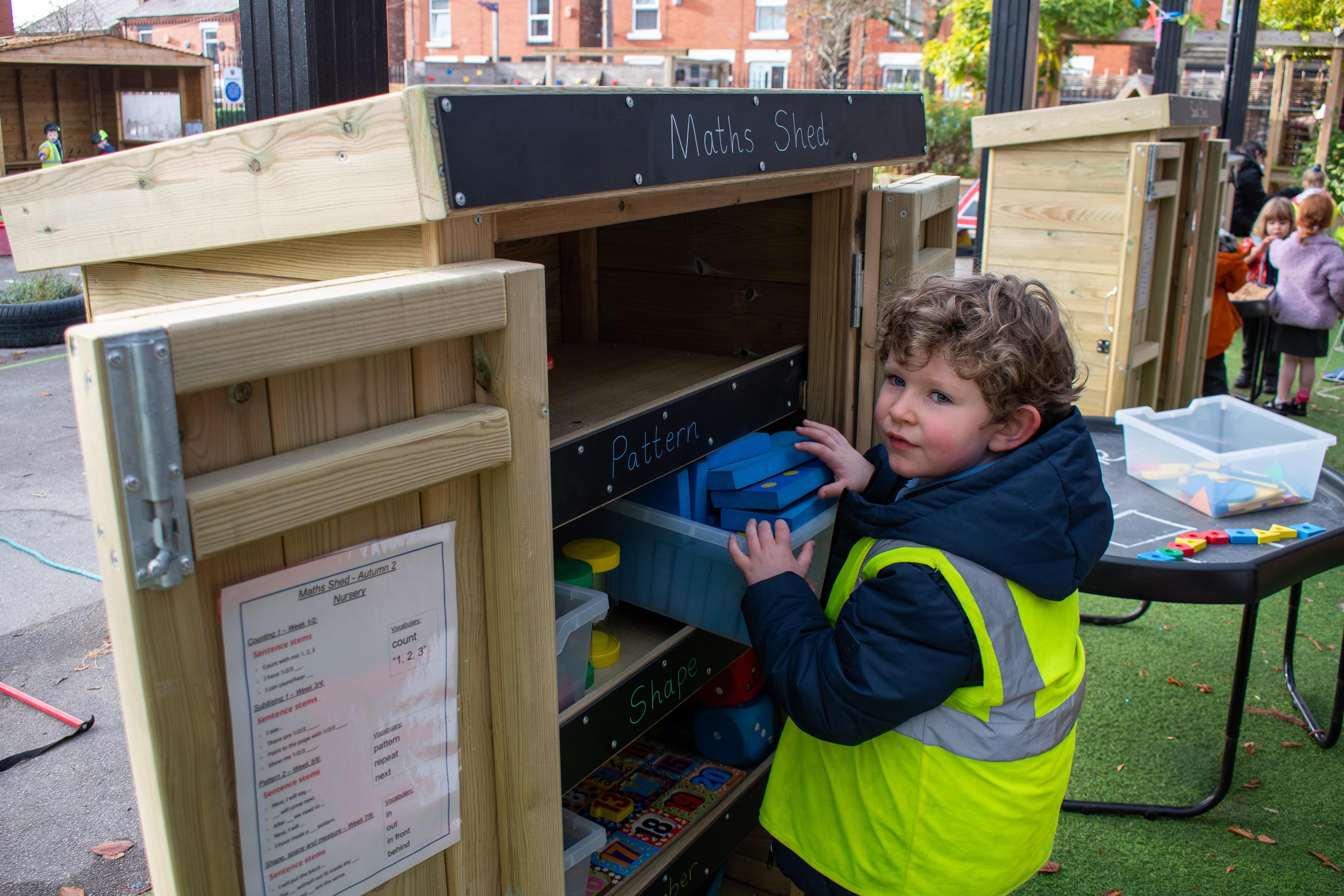 A child, who is wearing a coat and a high-vis jacket, is taking a plastic box out of the Acorn Storage Unit.