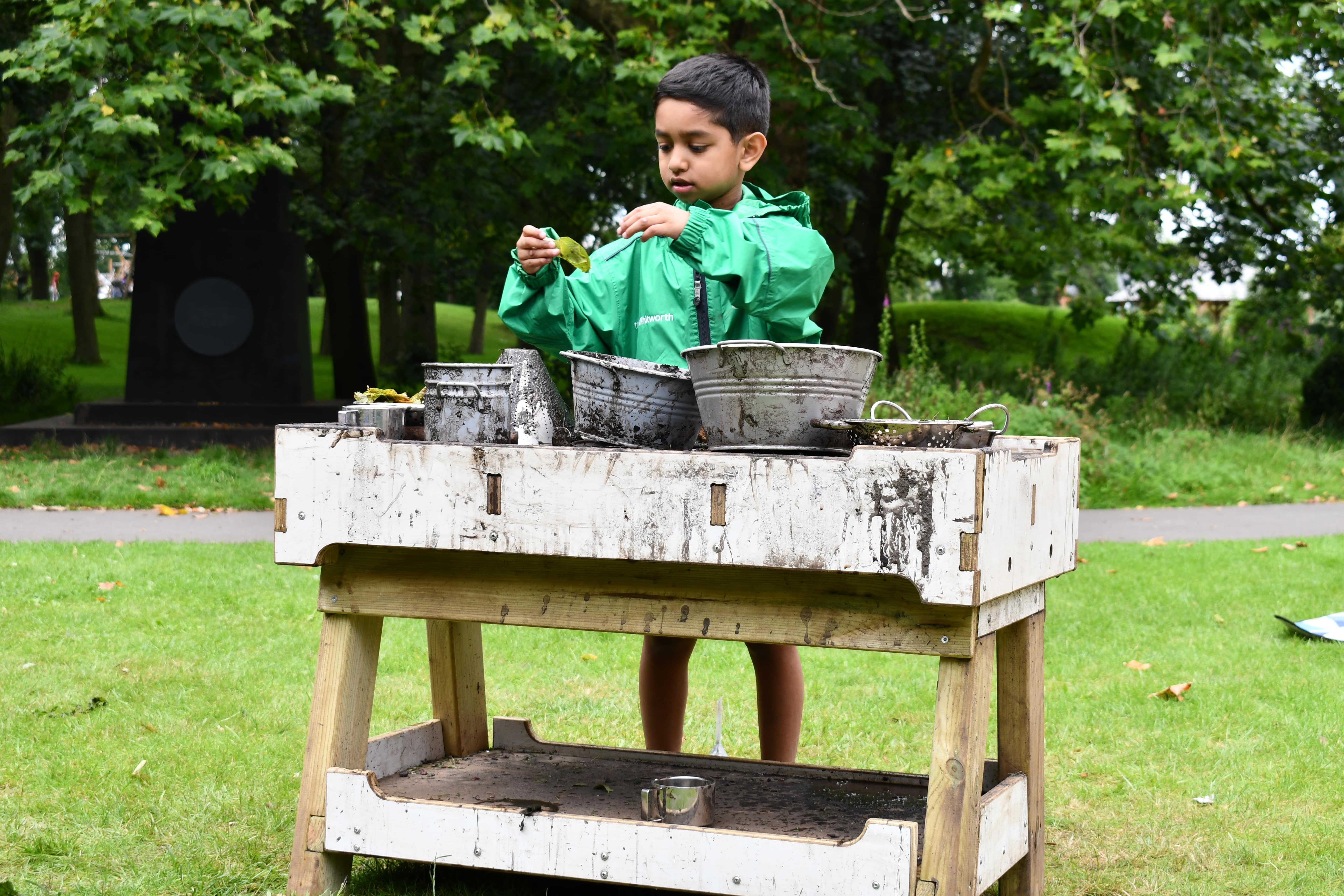 A little boy is wearing a green coat and is playing with a mud kitchen as it's raining.