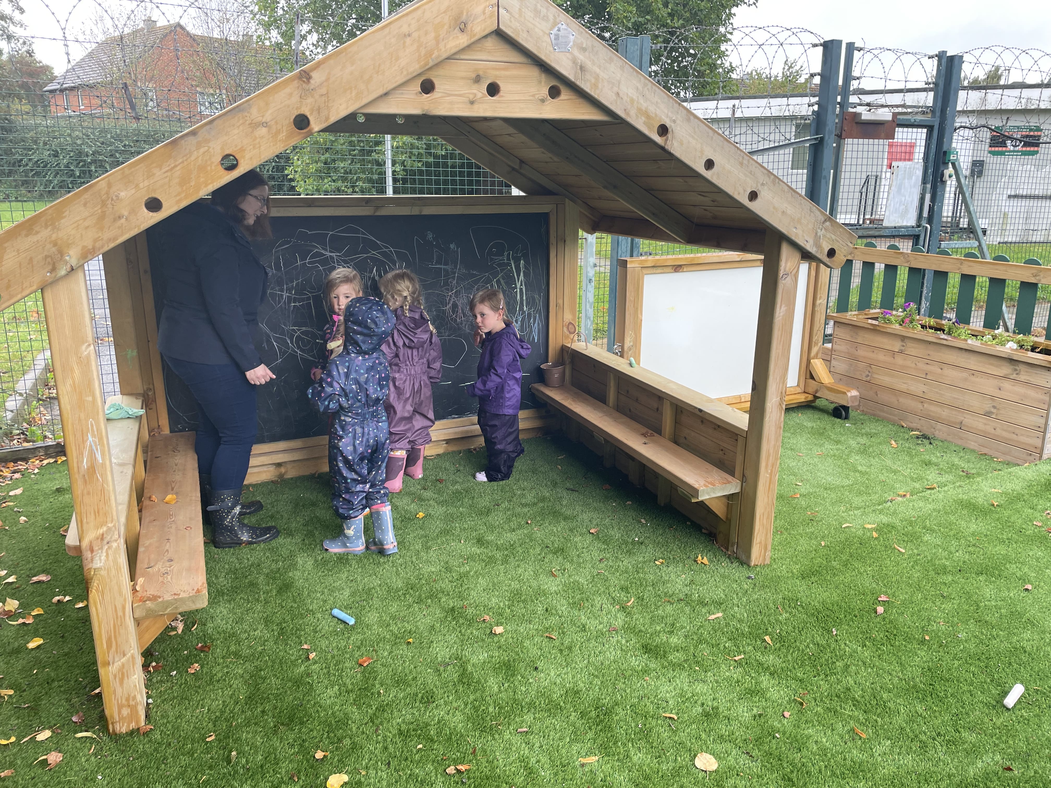 A group of 4 children are stood underneath a Giant Playhouse, as they draw on the chalkboard on the back wall. A teacher is helping them draw.