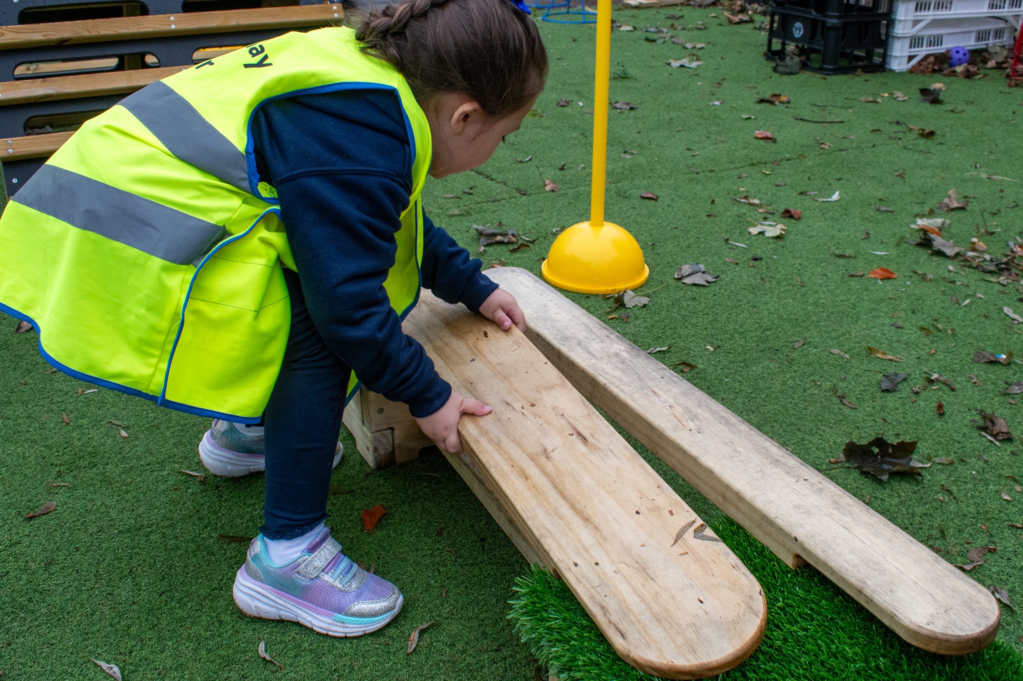 A little girl is wearing a high-vis jacket as she picks up a wooden plank which is covered in rain. The planks are placed on top of artificial grass.